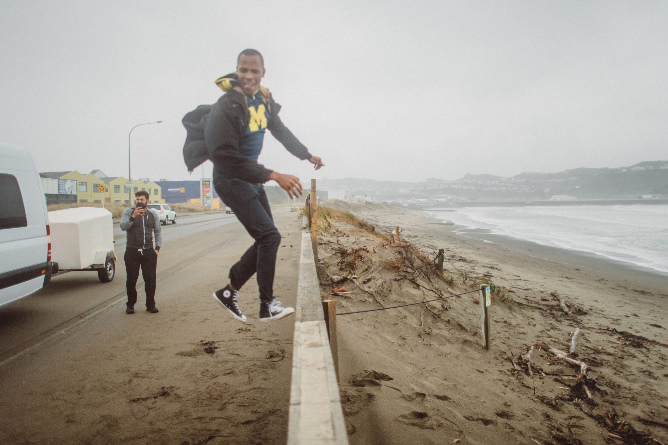 JSM African American man wearing a Michigan sweater jumping next to a fence by a beach with another man in the background