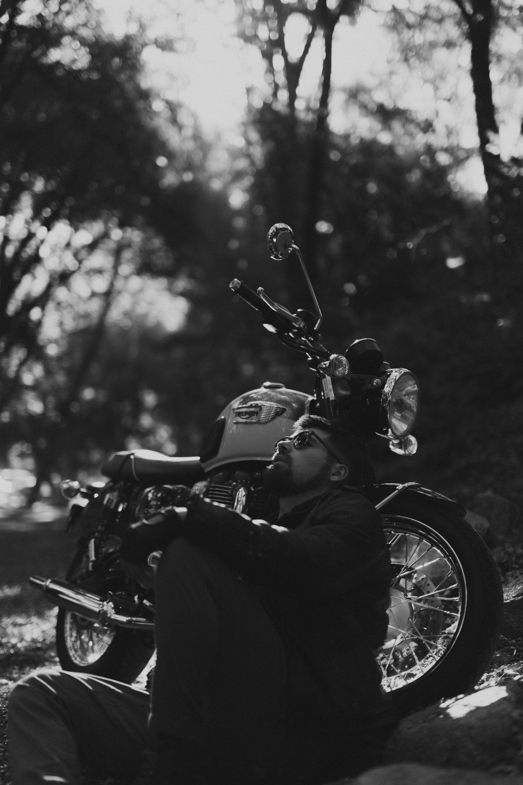 IU CI Studios Portfolio Triumph Motorcycles Euro Bike Shoot Black and white of motorcycle rider wearing shades relaxing sitting near the motorcycle posing for the camera and looking up at the sky