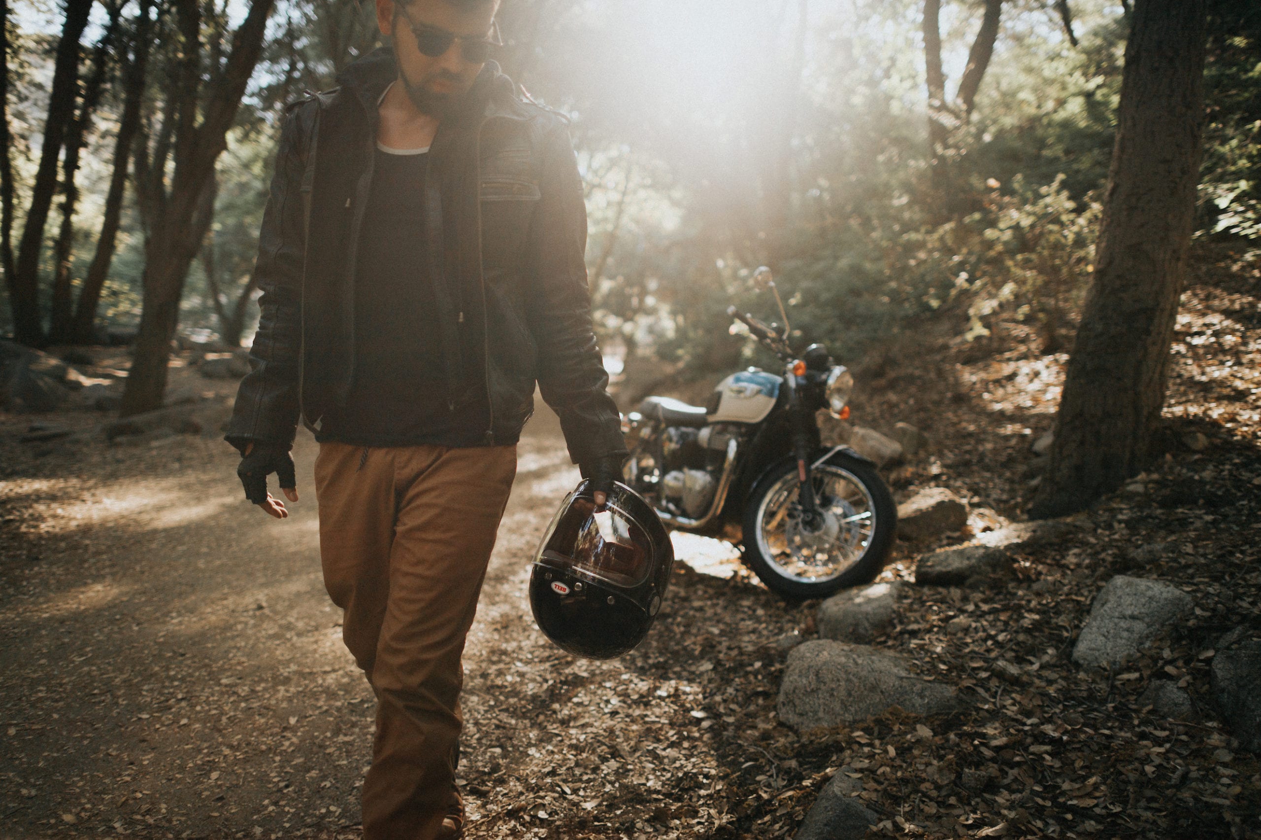 IU CI Studios Portfolio Triumph Motorcycles Euro Bike Photography Motorcycle rider wearing shades and leather gear poses for the camera in the middle of the forest holding his Bell helmet