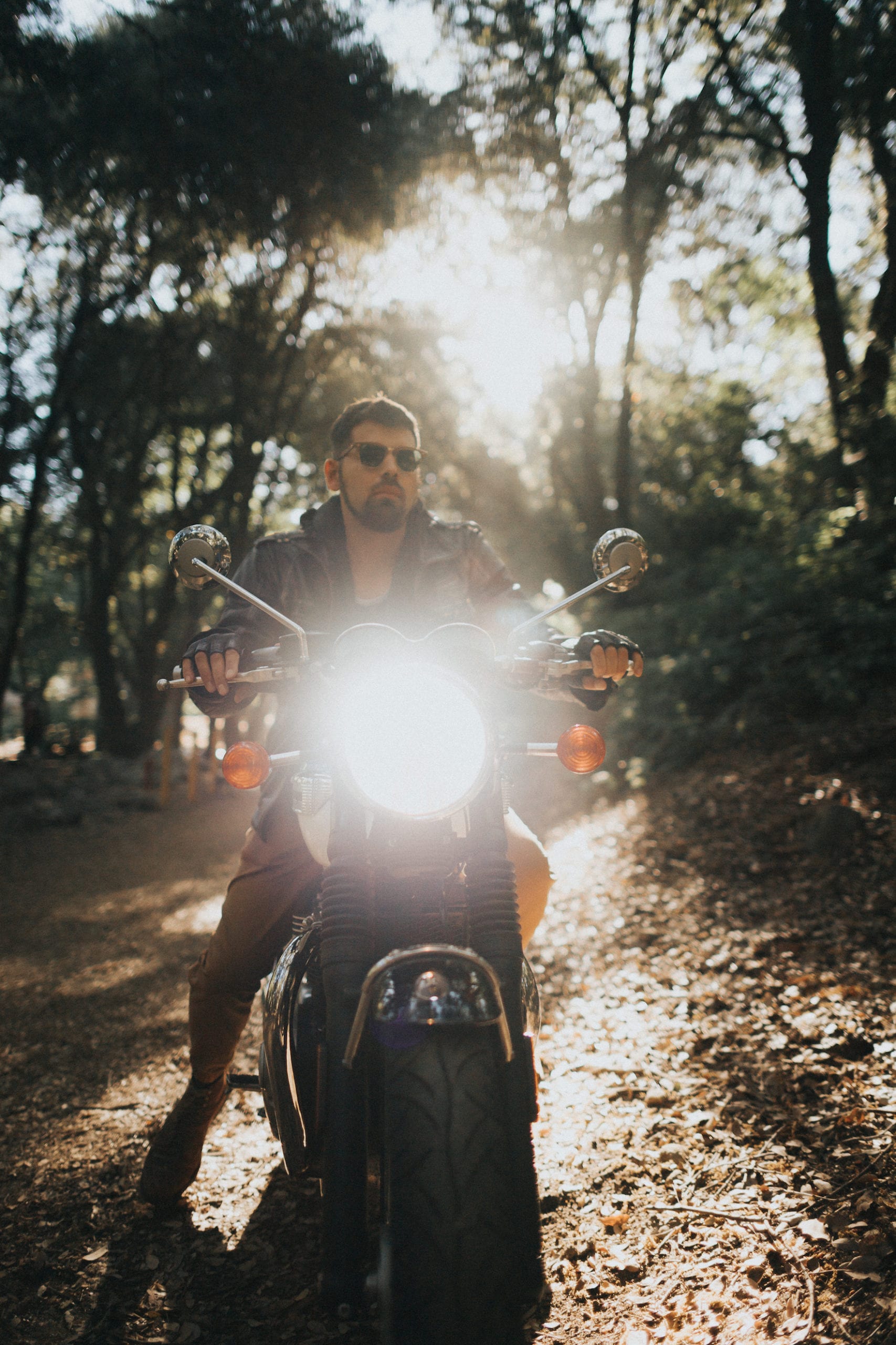 Triumph Motorcycles Euro Bike Photography Motorcycle rider wearing shades poses in the middle of the forest, pointing a shining headlight towards the camera