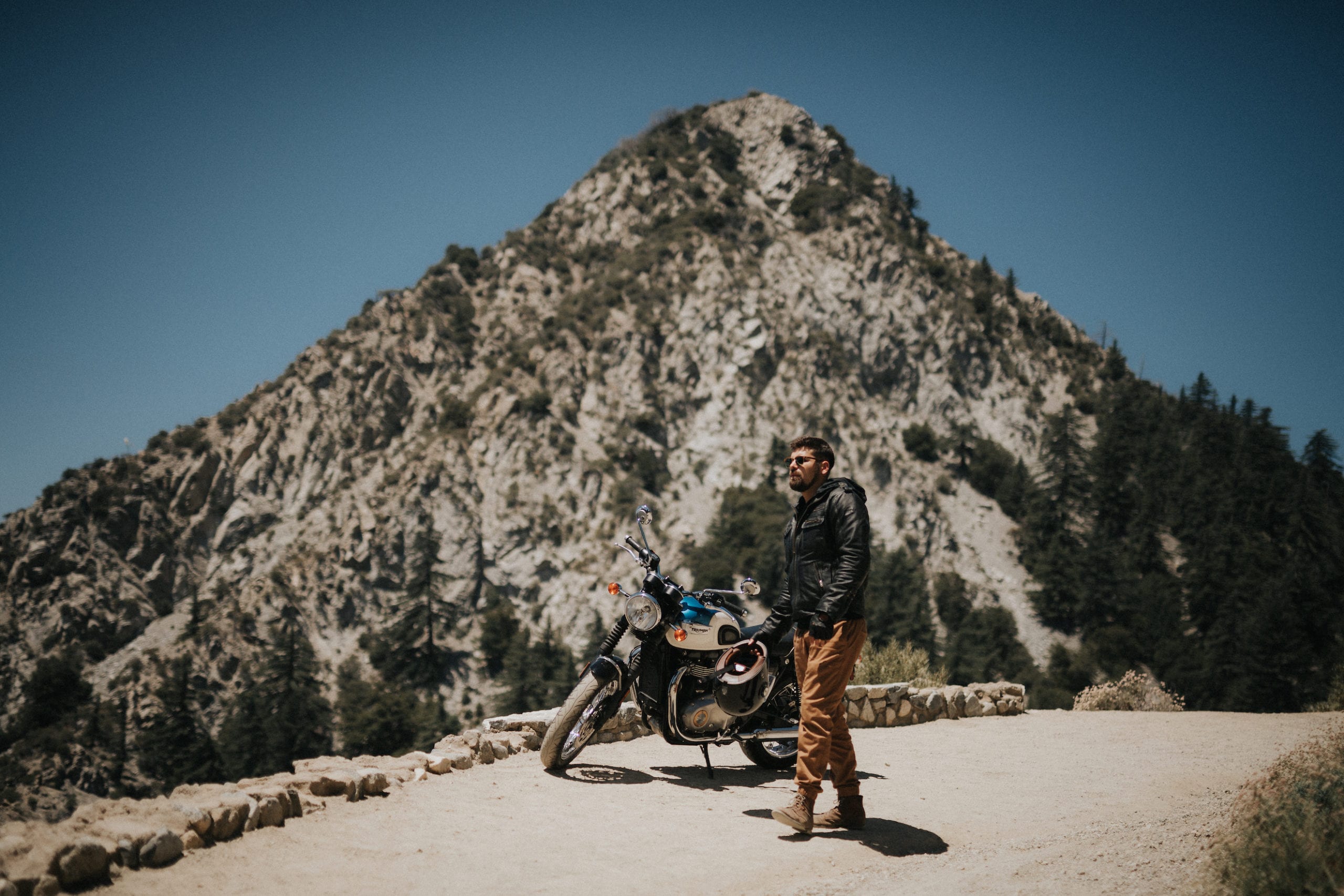 Triumph Motorcycles Euro Bike Shoot Motorcycle rider standing in front of a Bonneville T100 motorcycle with mountain peak in the backdrop