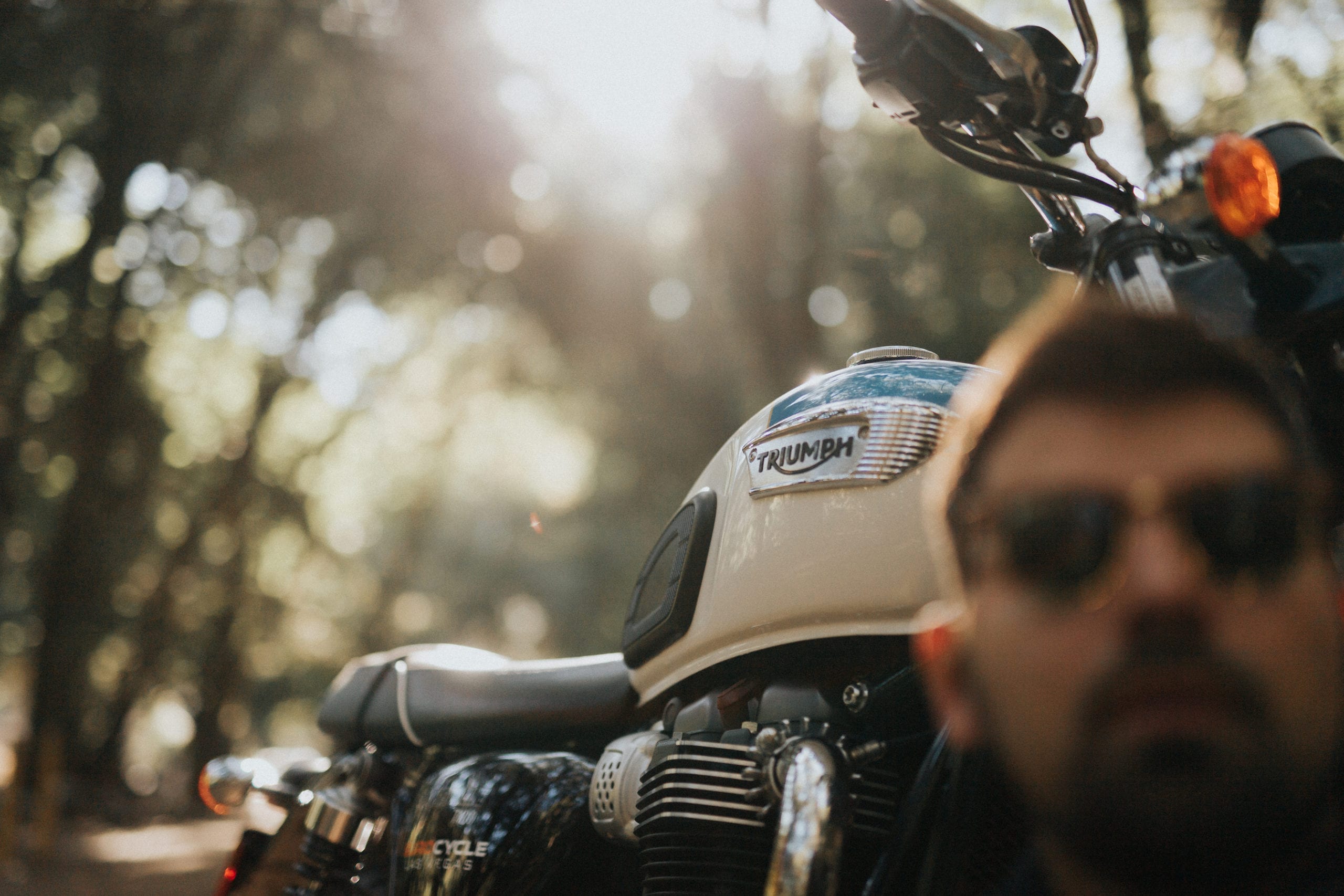 Closeup of motorcycle with rider in the foreground sitting in front of it wearing shades