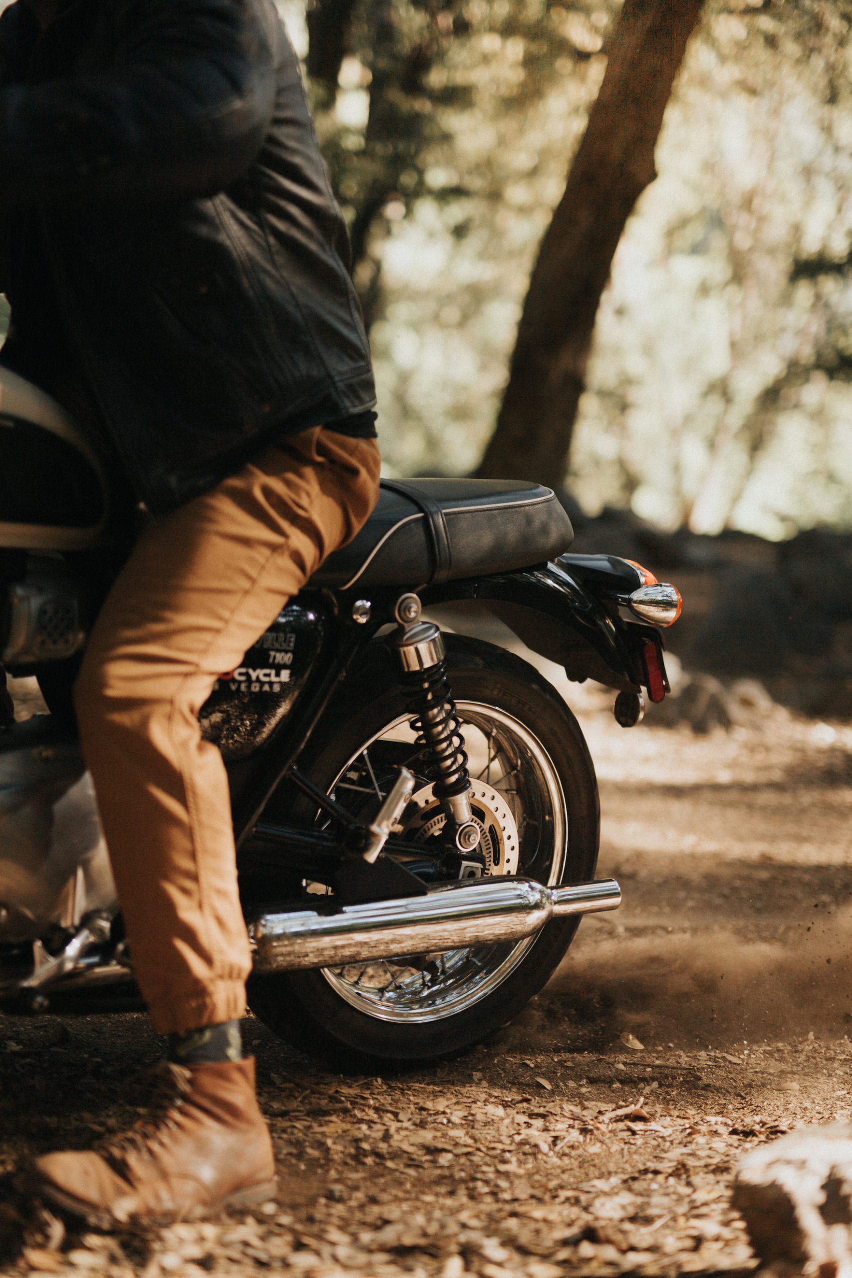 Closeup of backside of motorcycle with lower half of man in leather boots along with burnt orange pants and black leather jacket sitting on it