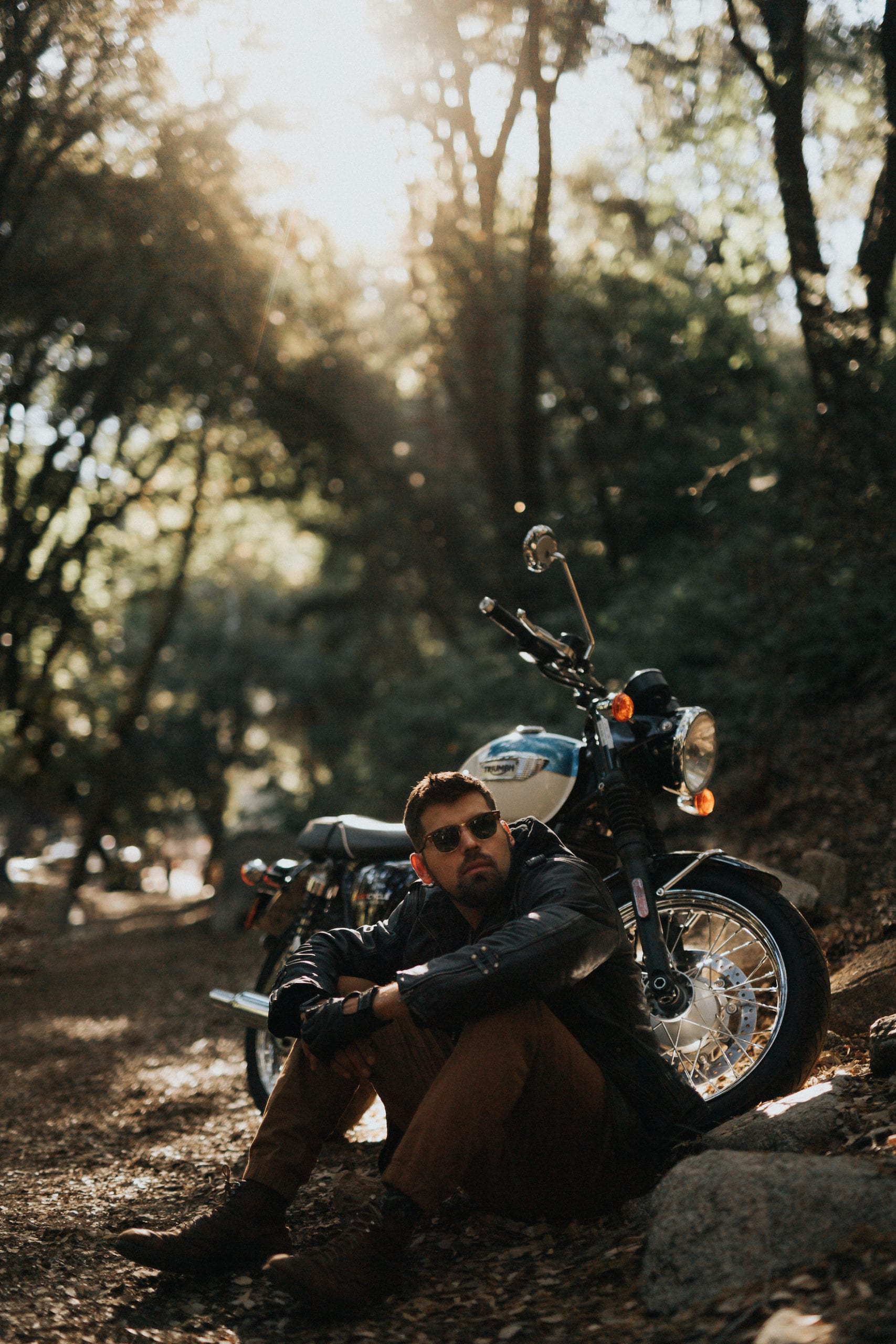 Motorcycle rider relaxes sitting in leaves near the motorcycle posing for the camera and looking off to the side