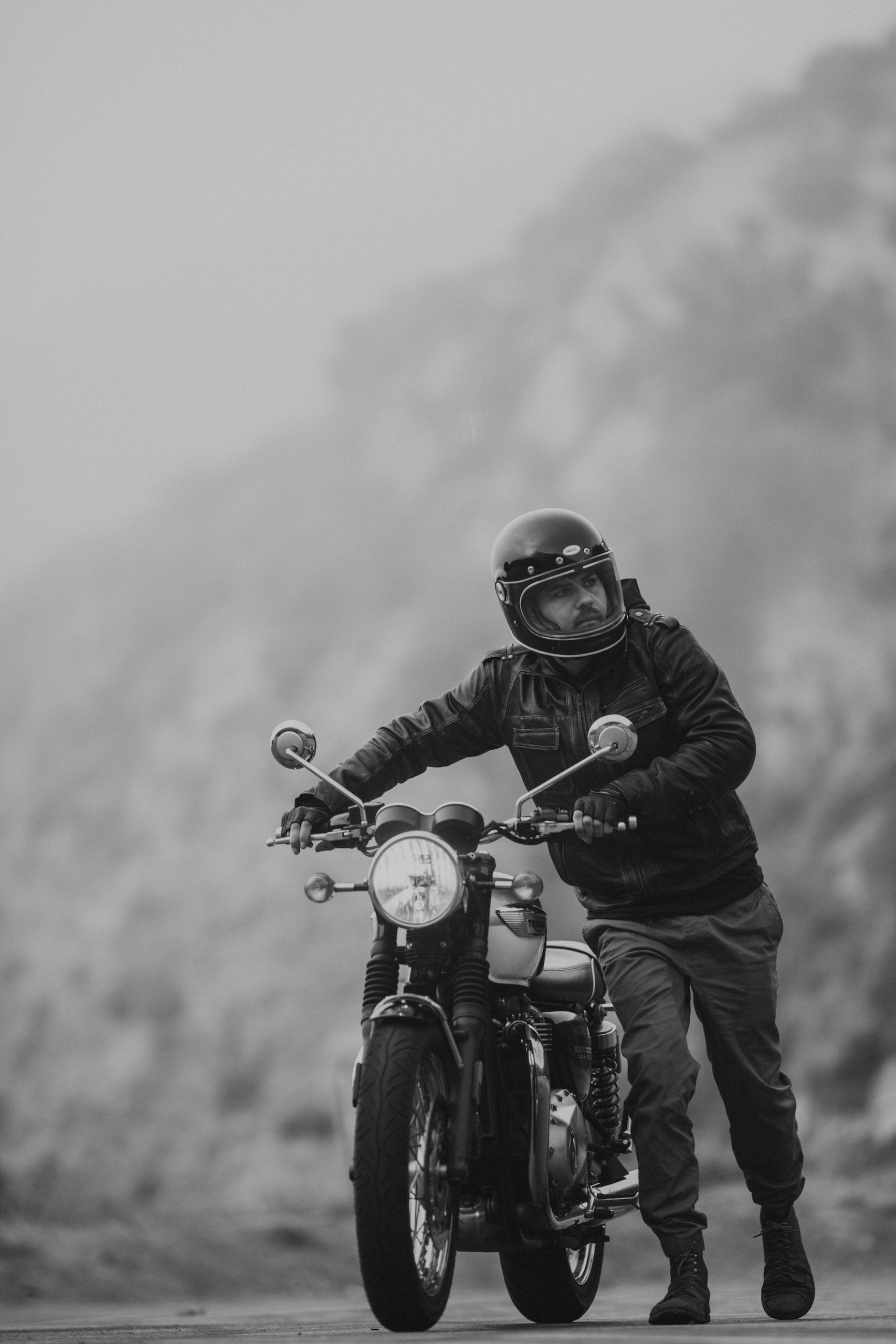 Motorcycle rider wearing helmet and leather motorcycle gear pushing the Bonneville T100 up a bend on the side of the road.