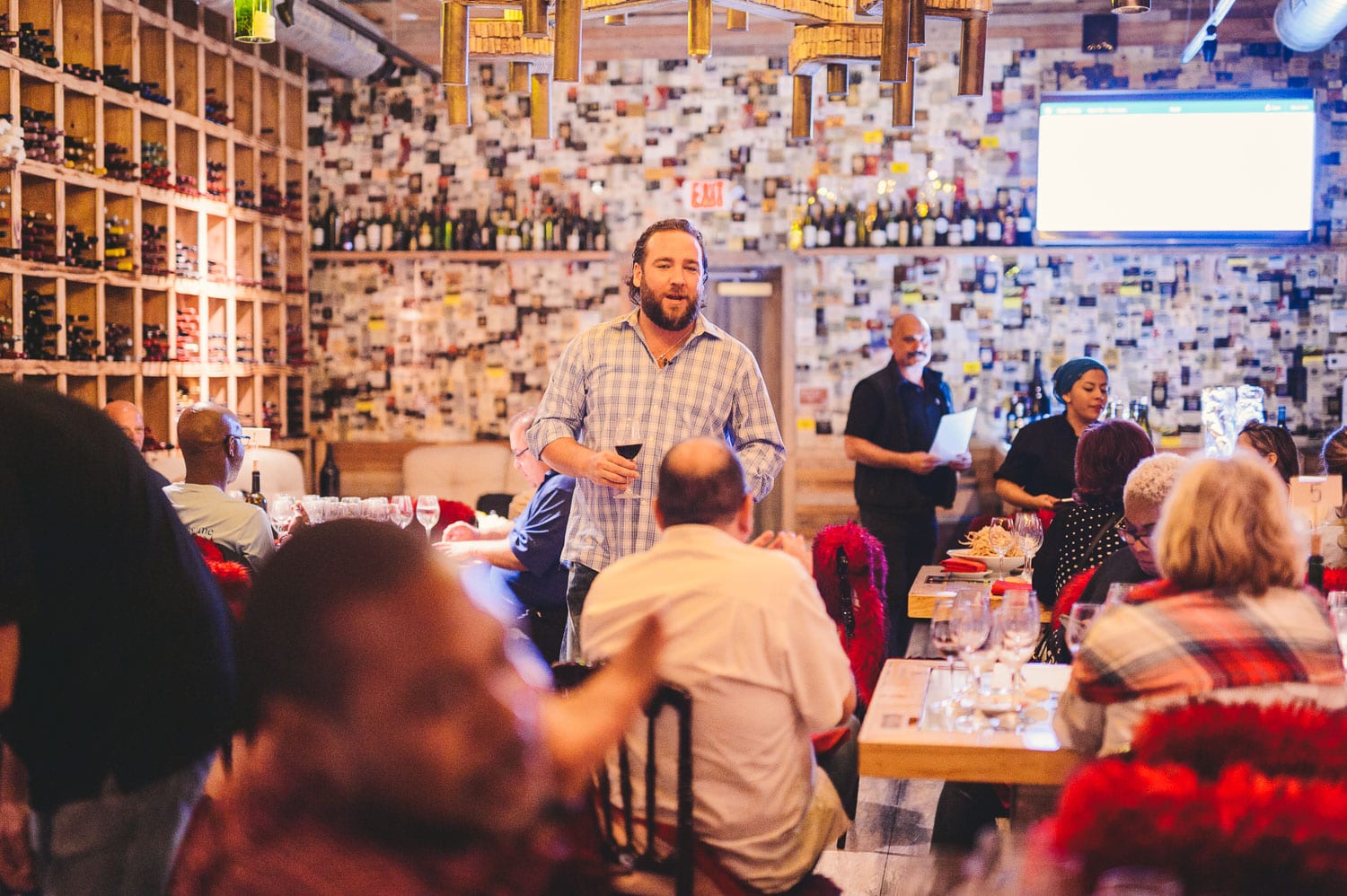 Misc image Man wearing a blue squared shirt talking to a group of people in a wine cellar holding a glass of wine