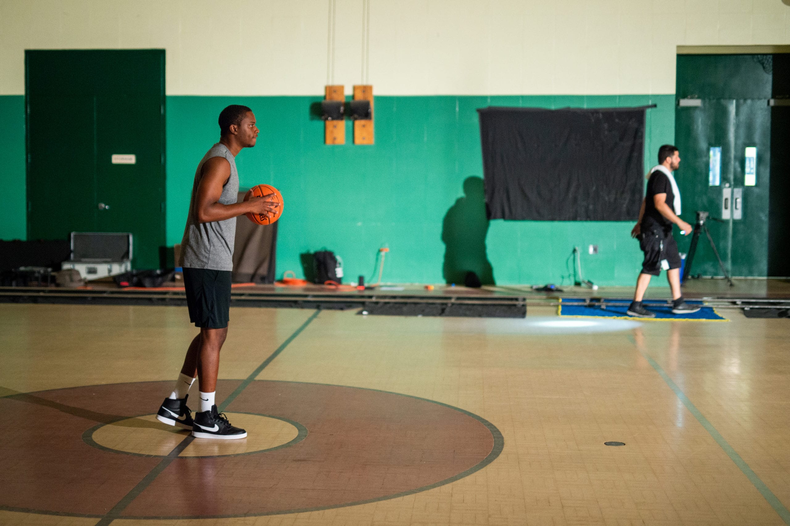 Nike African American man wearing a gray t shirt holding a ball on a basketball court posing for the camera with a crew member walking off screen in the background