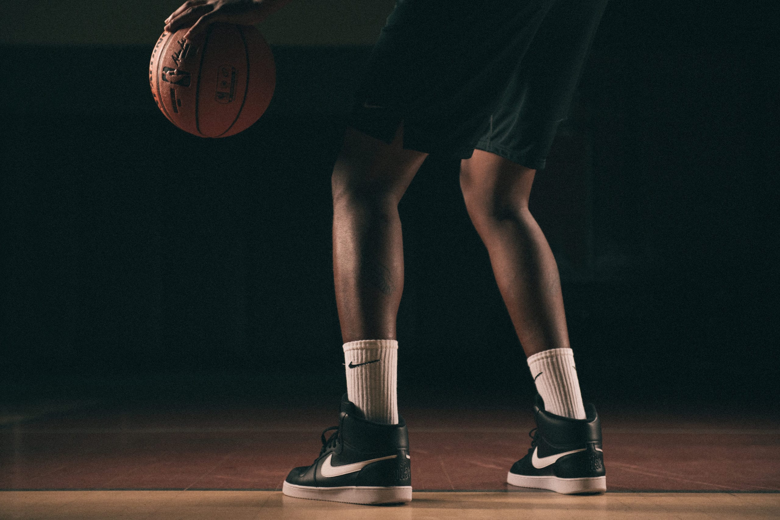 Nike Creative Marketing Concepts View from behind of the lower half of an African American man wearing black Nike shoes and white Nike socks poised holding a basketball on a basketball court with a light shining on him in semi darkness