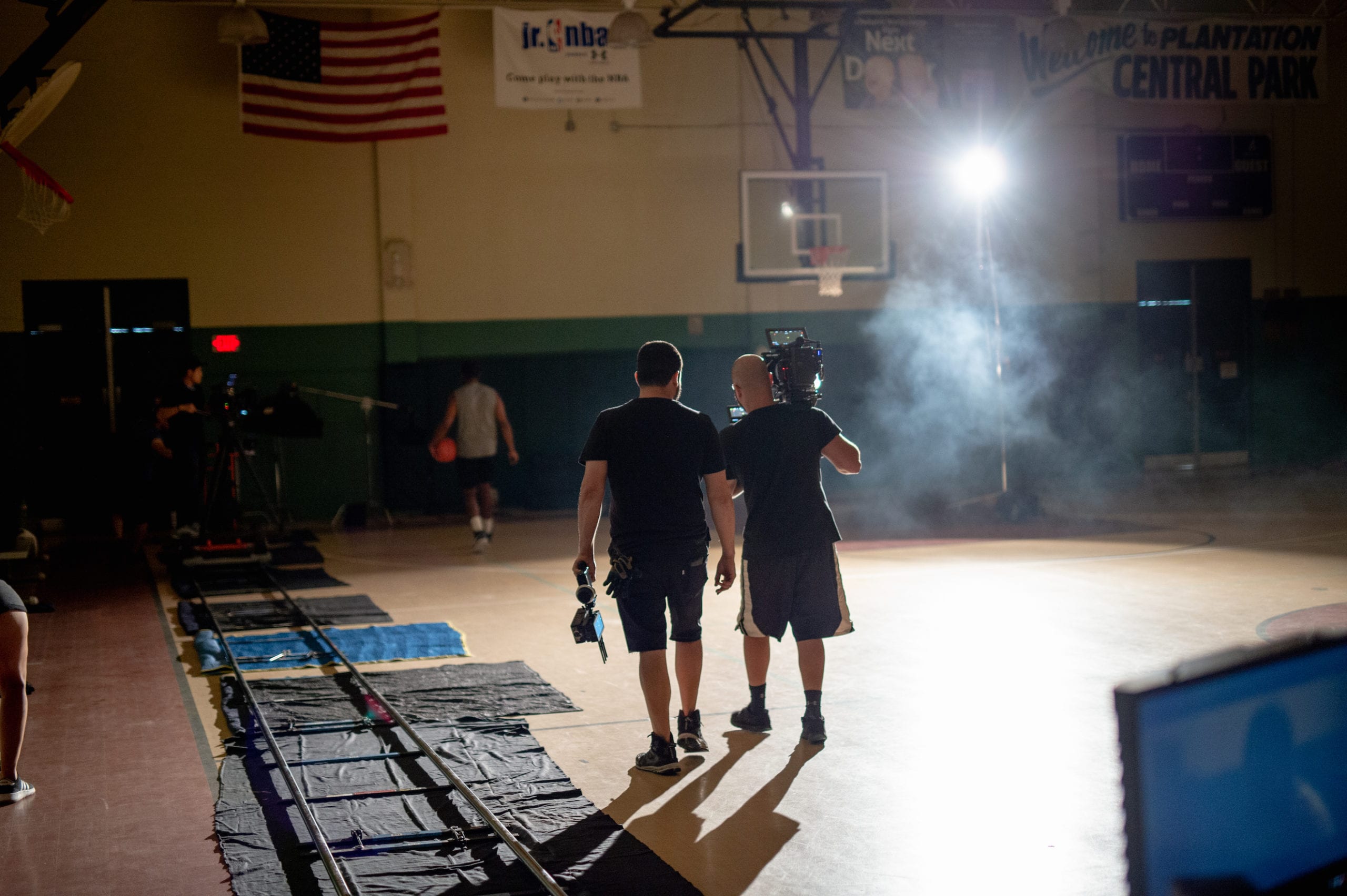 Nike scene being filmed on a basketball court in a gym with an African American man wearing a gray t shirt holding a ball walking away on the court surrounded by a film crew