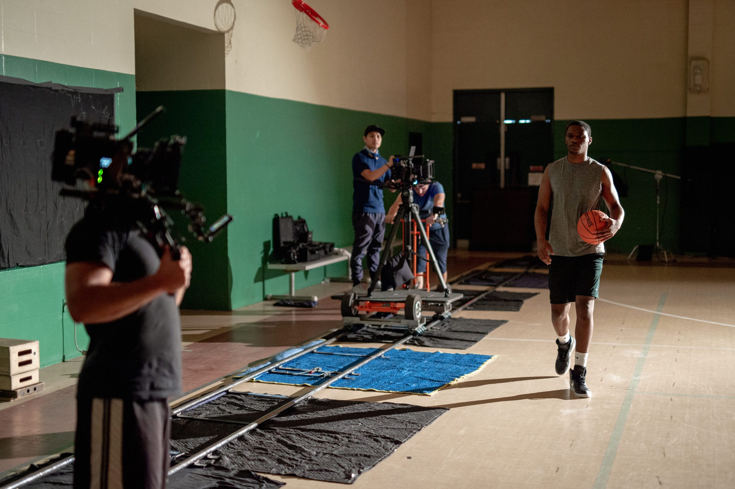 Nike scene being filmed on a basketball court in a gym with an African American man wearing a gray t shirt holding a ball walking on the court surrounded by a film crew