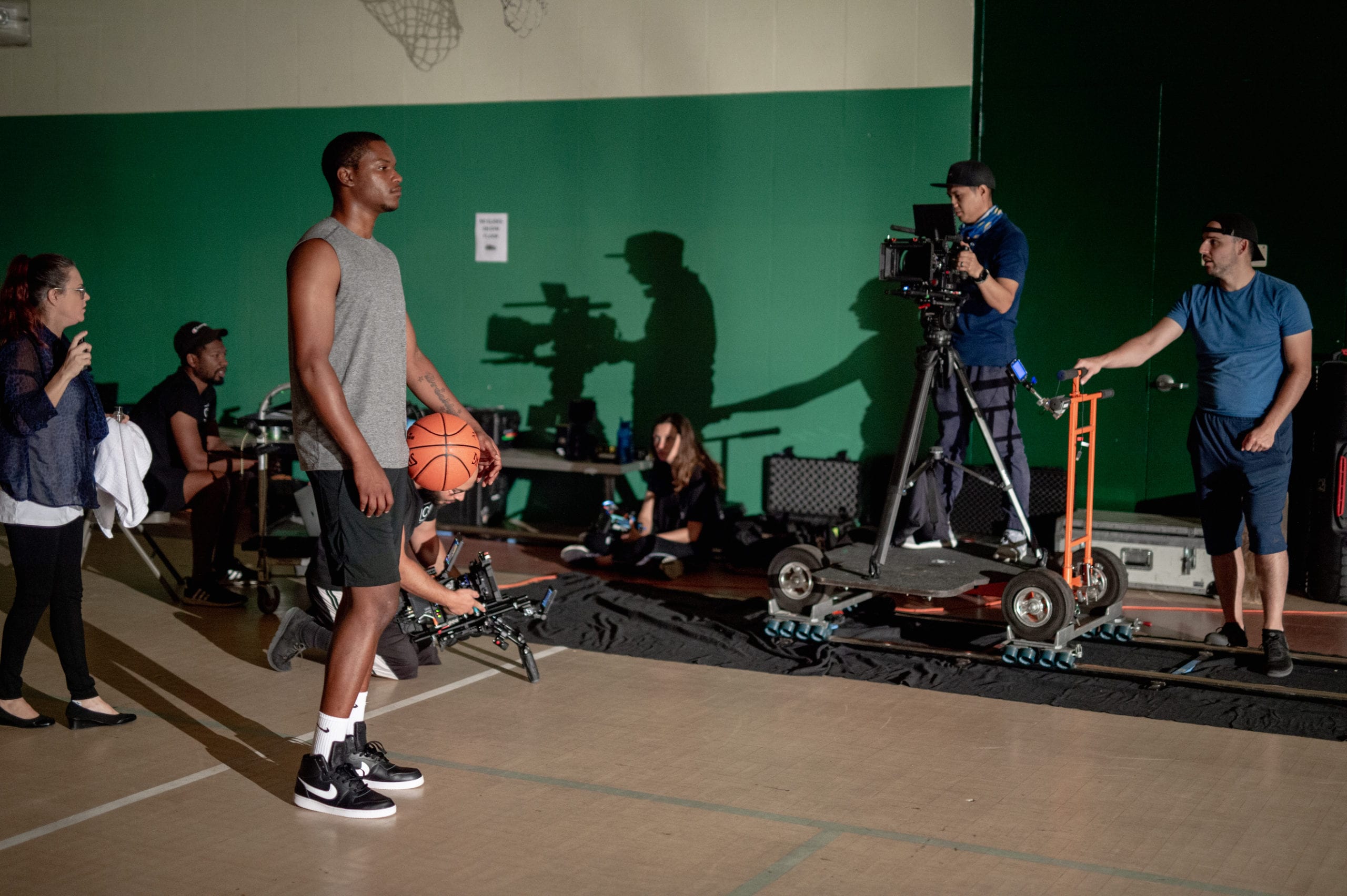 Video Production Behind the Scene being filmed on a basketball court in a gym with an African American man wearing a gray t shirt holding a ball surrounded by a film crew