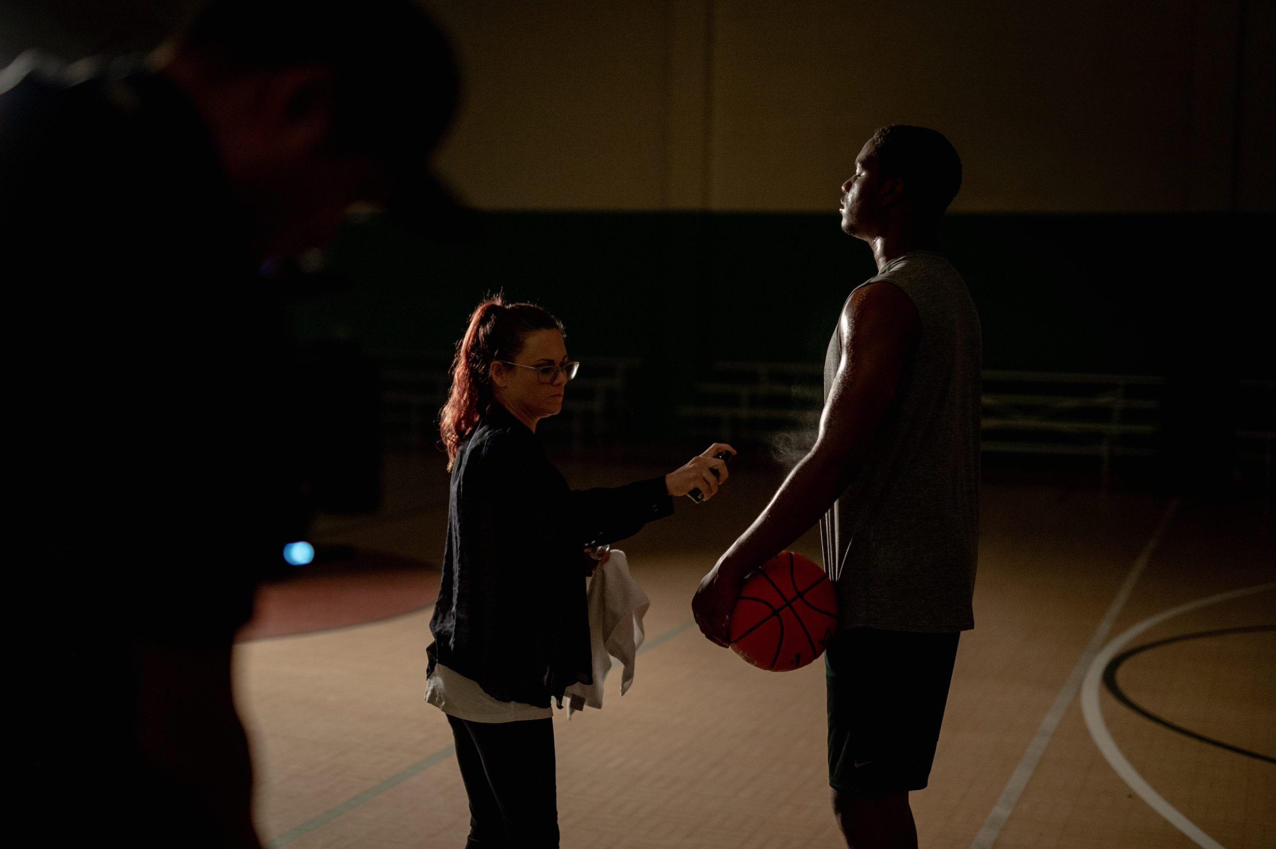 Nike scene being filmed on a basketball court in a gym with an African American man wearing a gray t shirt holding a ball being tended to by a makeup artist