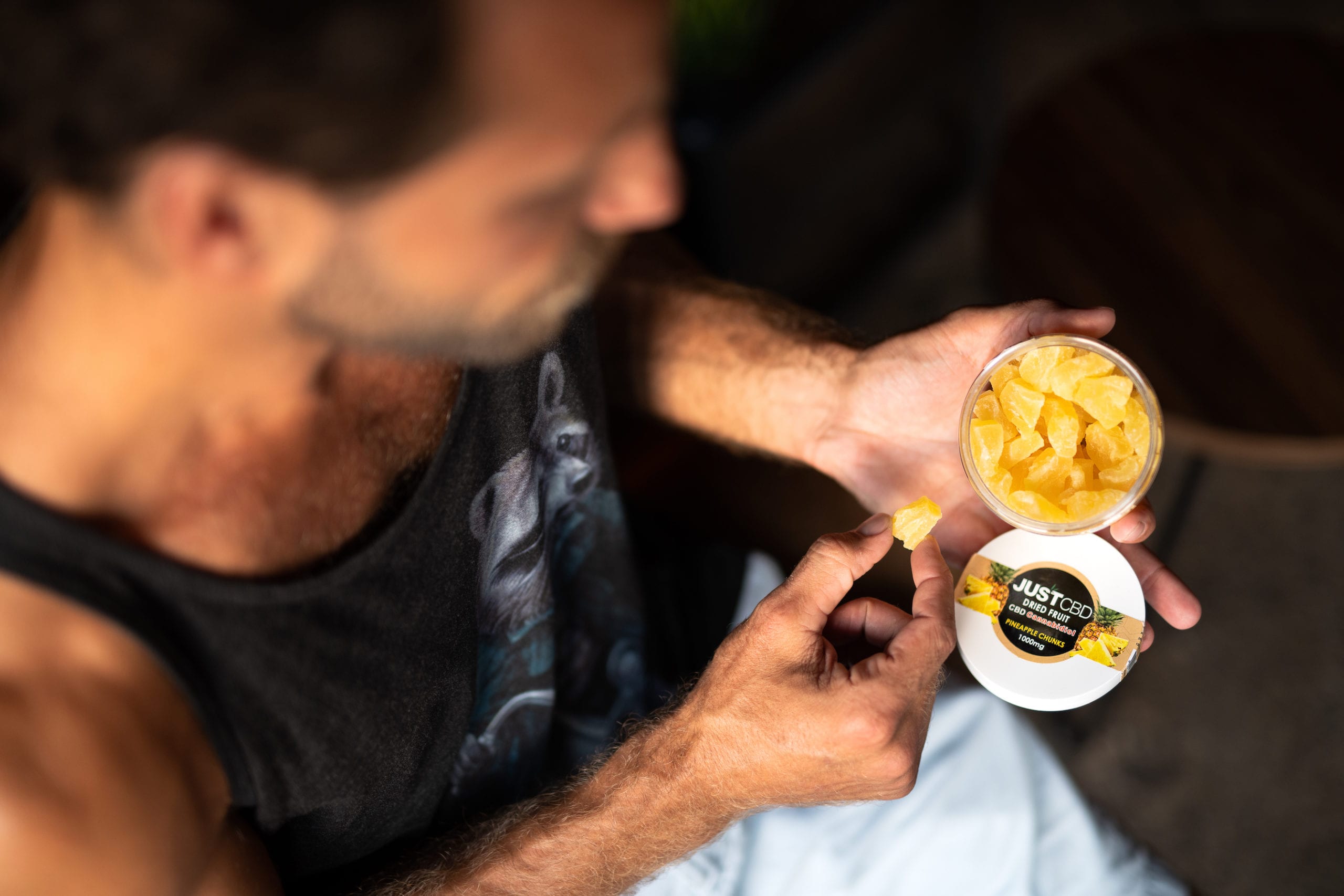 JustCBD Products and JustCBD Product Photography Overhead view of a man taking a pineapple chunk from JUSTCPD dried fruit container