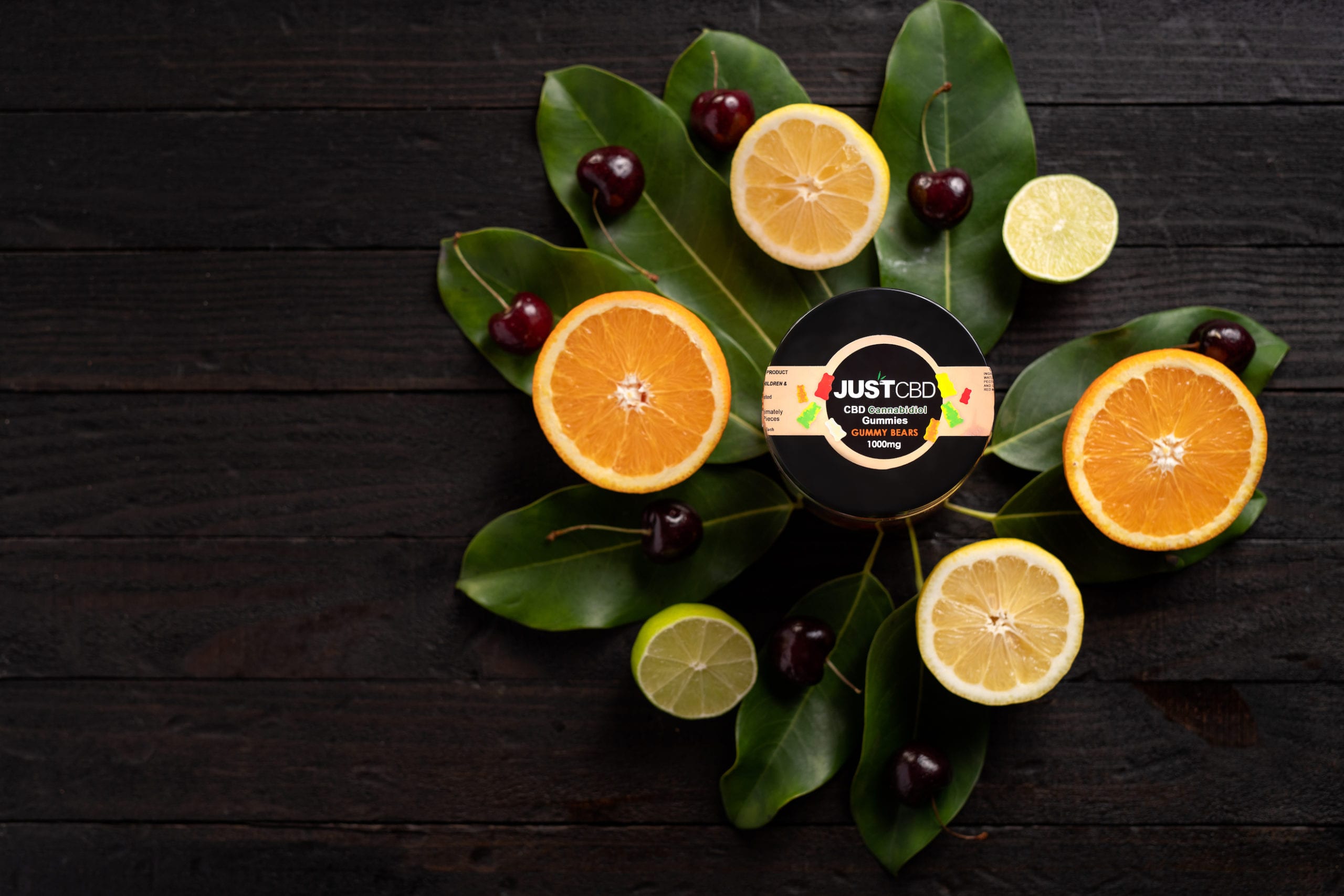 JustCBD Product Photography and Just CBD Products Overhead view of JUSTCBD gummy bears container on display surrounded by sliced citrus fruit and cherries on green leaves on a wooden table