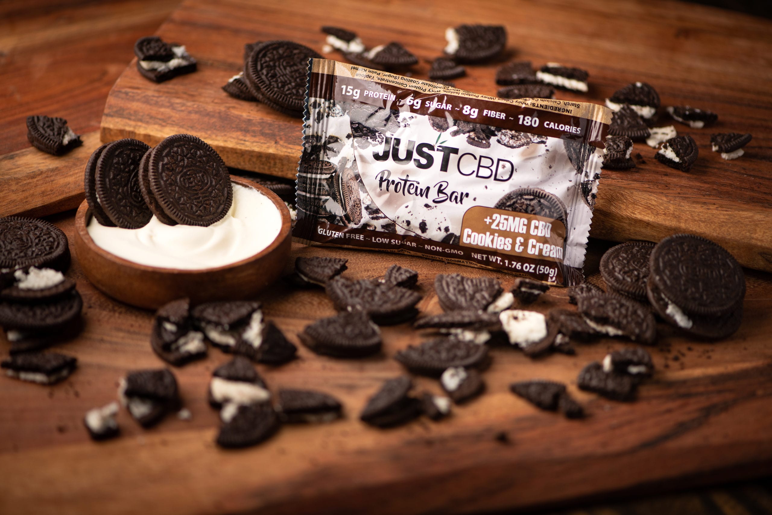 IU C&I Studios Portfolio JustCBD Product Photography and Just CBD Products JUSTCBD Cookies and cream protein bar package surrounded by cookies and bowl of cream on a wooden table