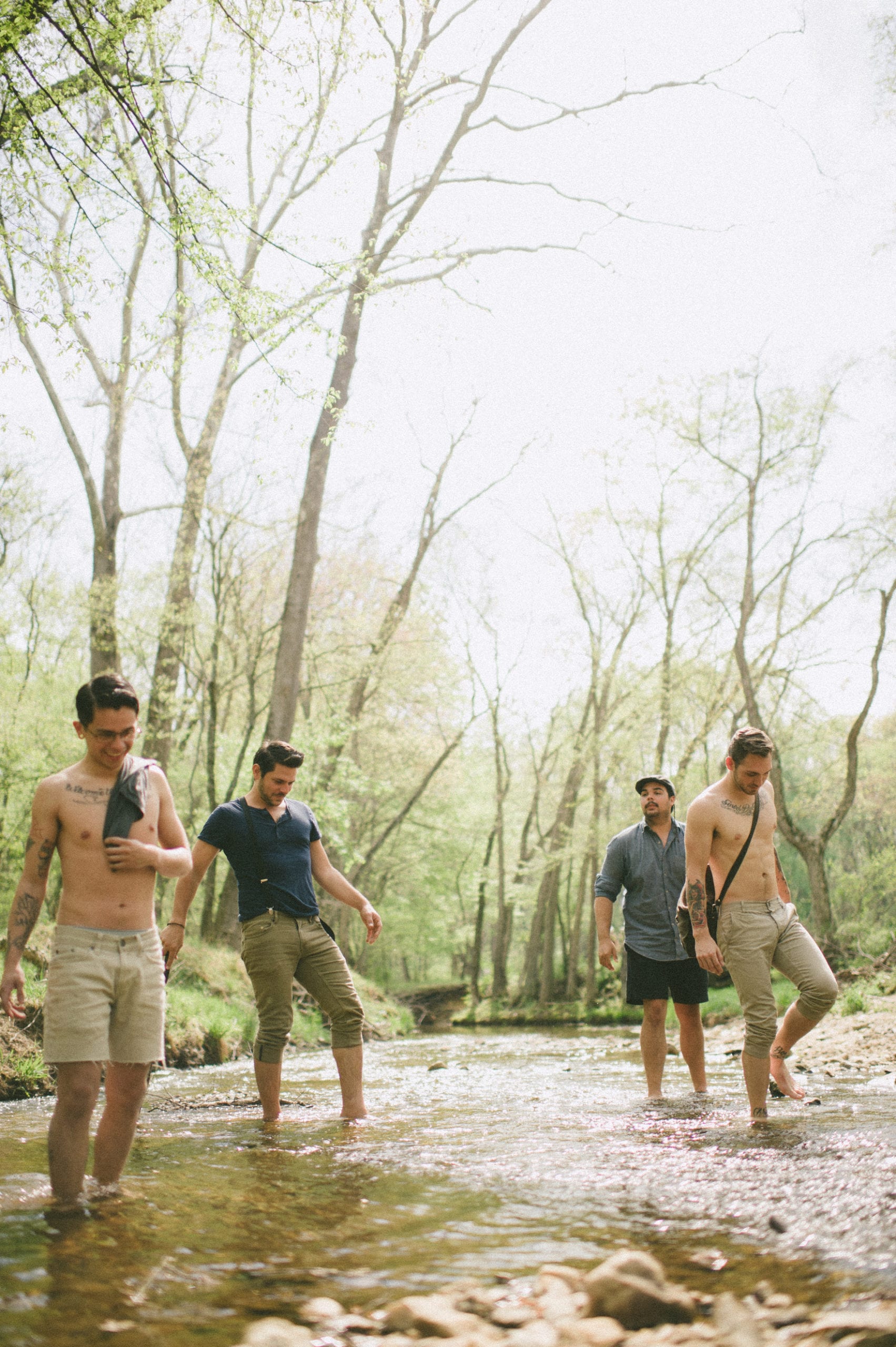 Kids American Indie Rock Band Four males walking in a creek on a sunny day in the forest. Two males are shirtless with tattoos on their arms and chest.