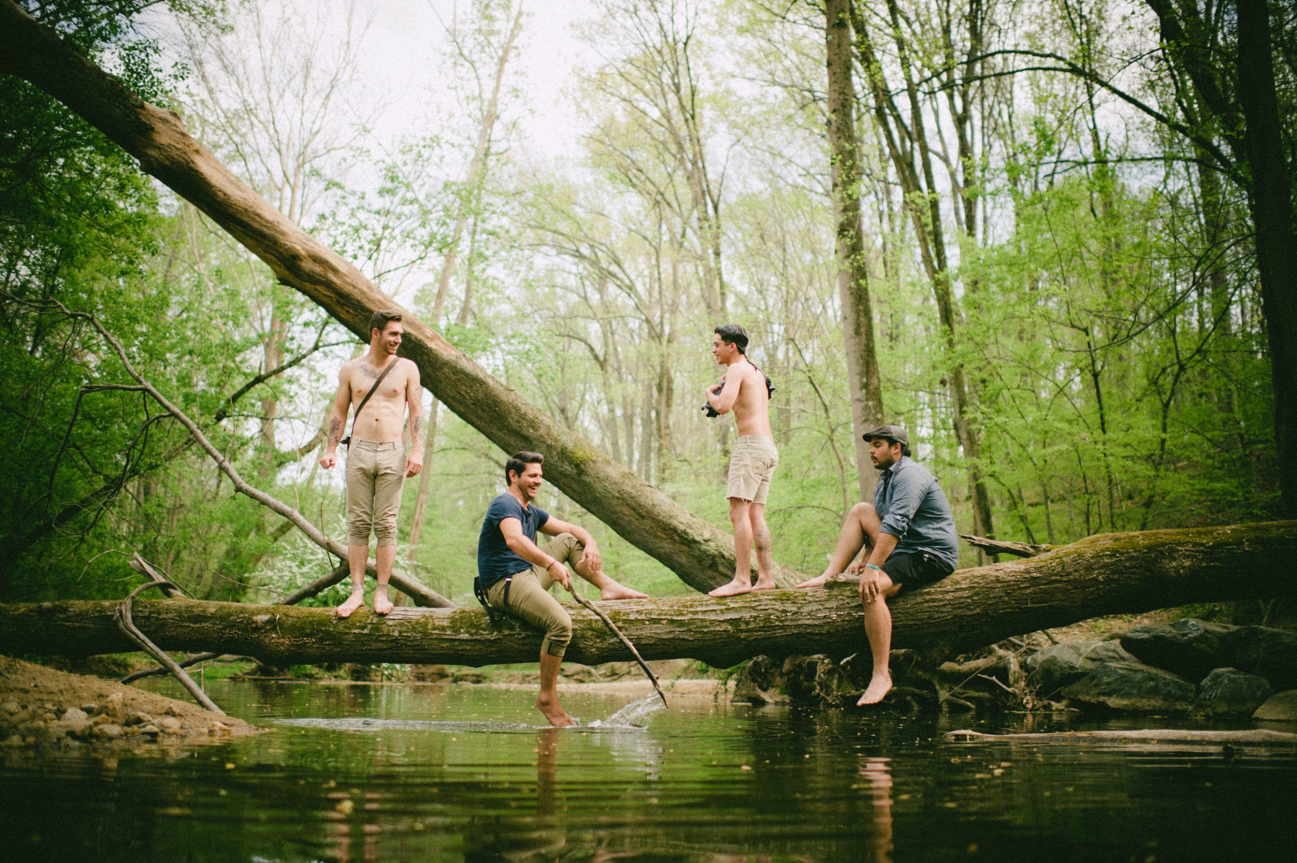 Kids American Indie Rock Band Four men in the forest by a creek. Two are shirtless and are standing on a log and two are sitting sideways on a log facing each other with one holding a stick.