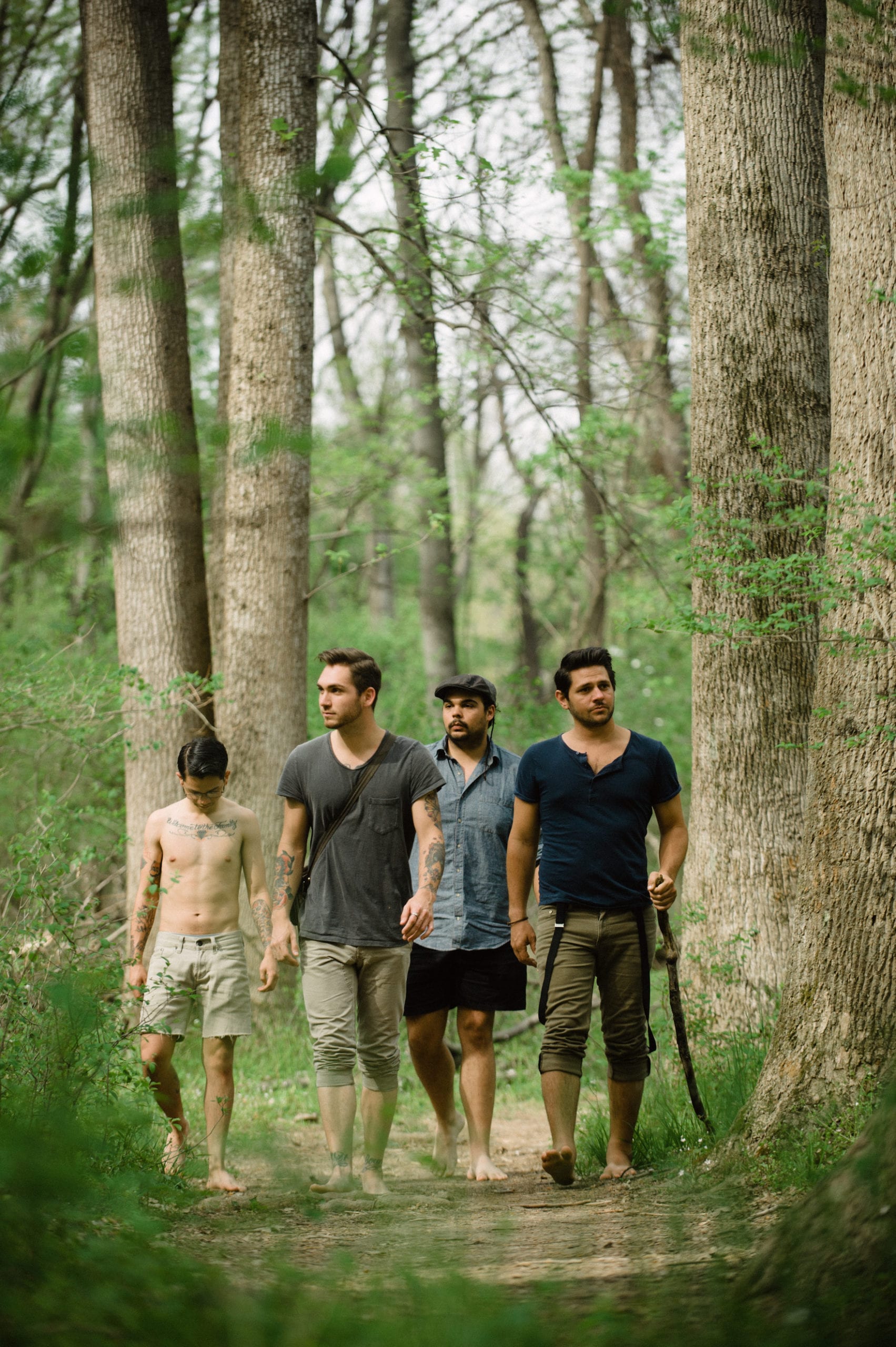 Kids American Indie Rock Band Four men walking in a forest with one shirtless with tattoos and one holding a stick
