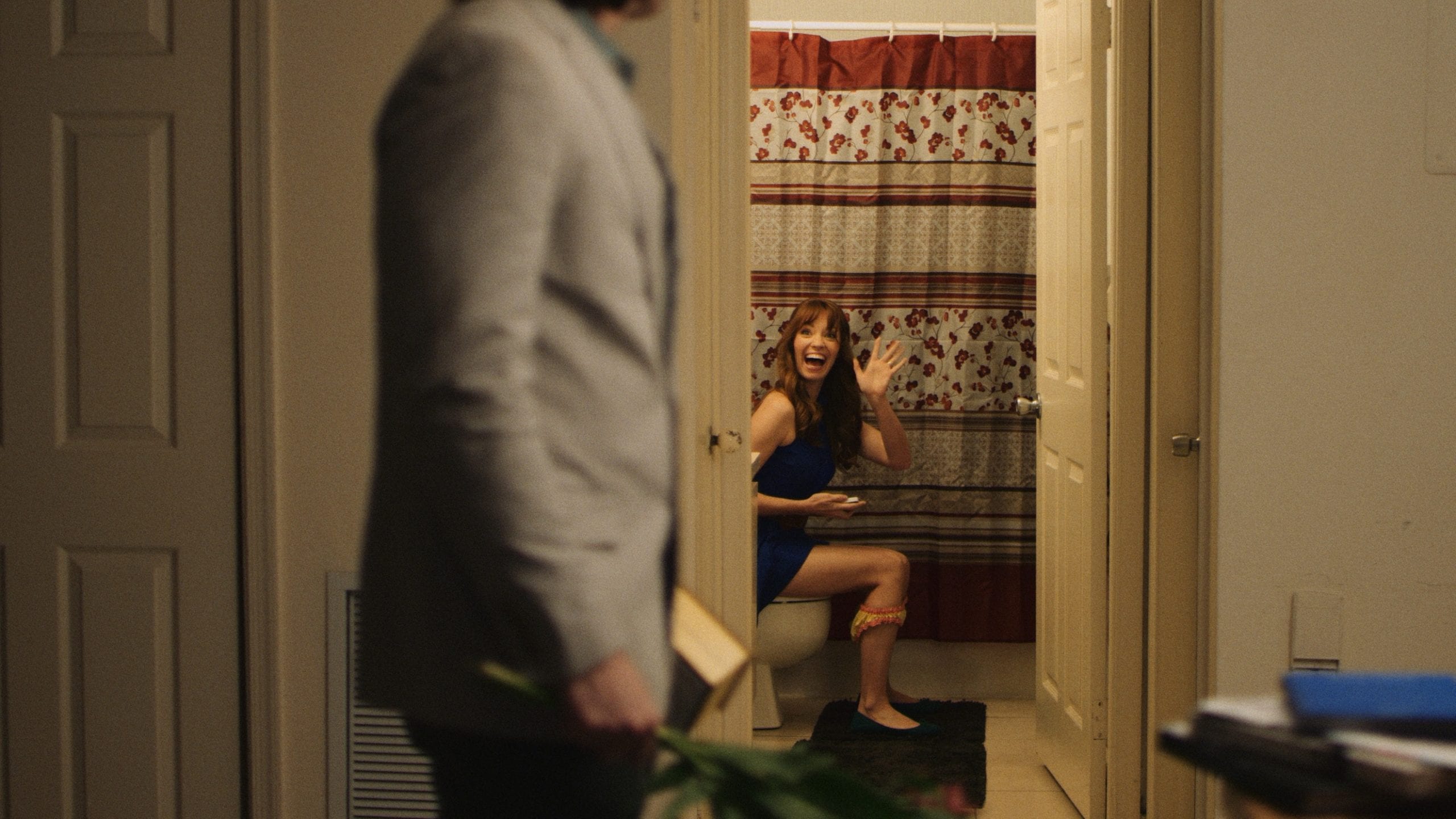 Poop Man in a gray suit carrying a box of Whitman's Chocolates and flowers walking past a bathroom with a woman sitting on the toilet waving and smiling