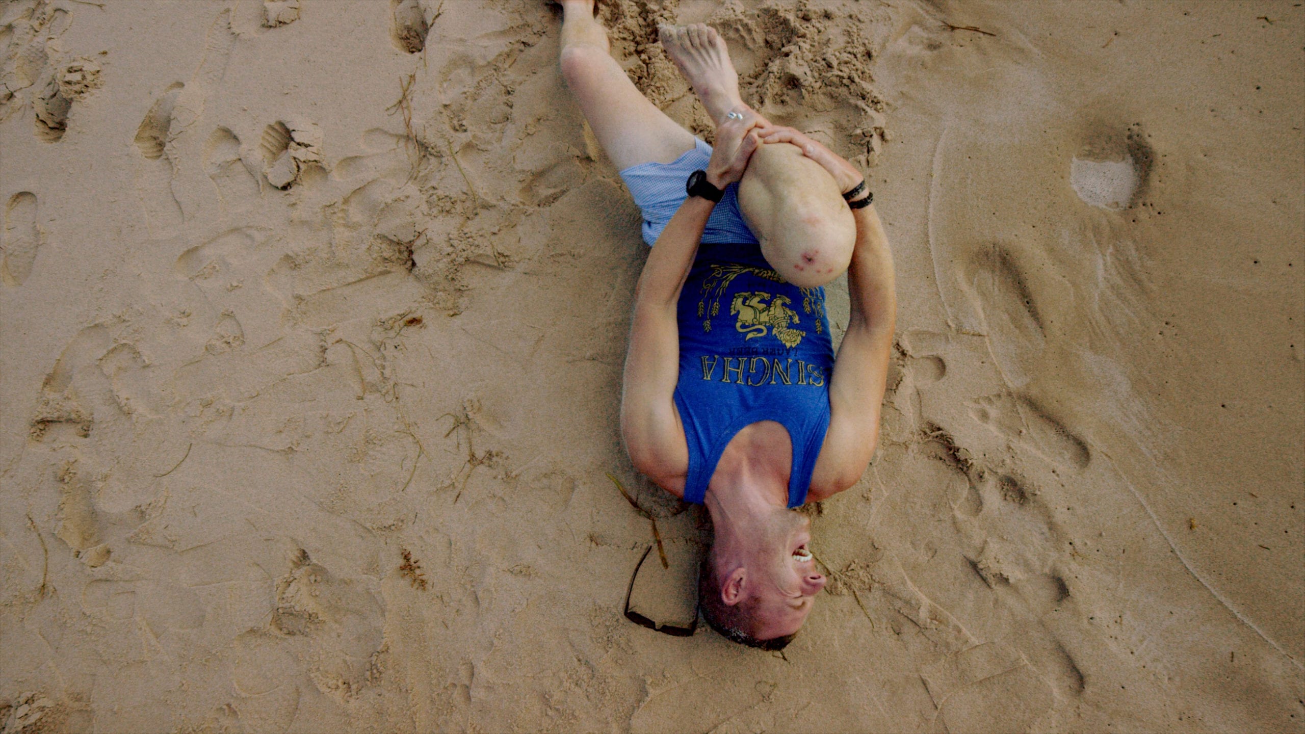 REVA Air Ambulance Overhead view of man wearing blue tank top laying in the sand clutching his leg with scraped knee grimacing in pain