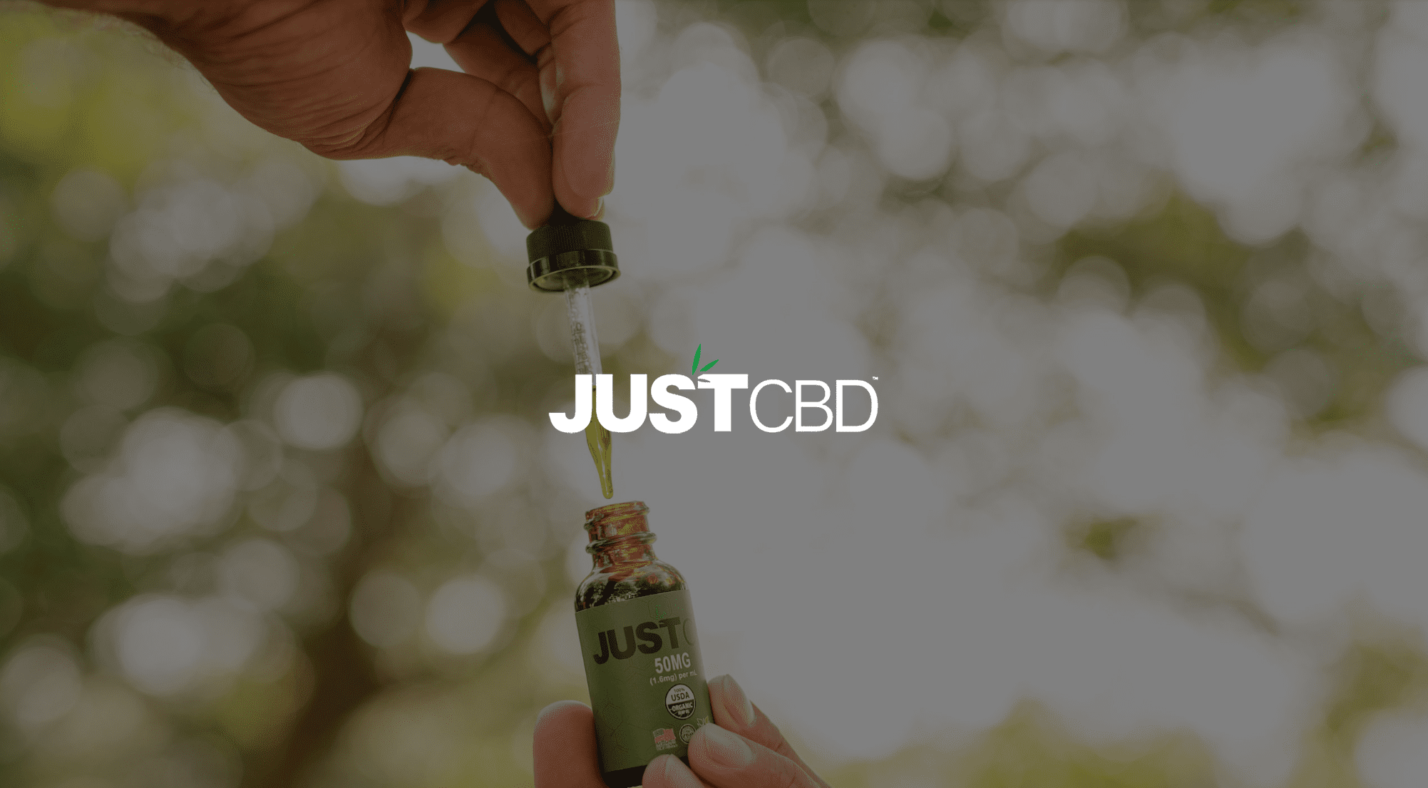 IU C&I Studios Page White JustCBD logo with green leaves with background of bottle of JUSTCBD Hemp Seed Oil being held with a filled dropper poised above it