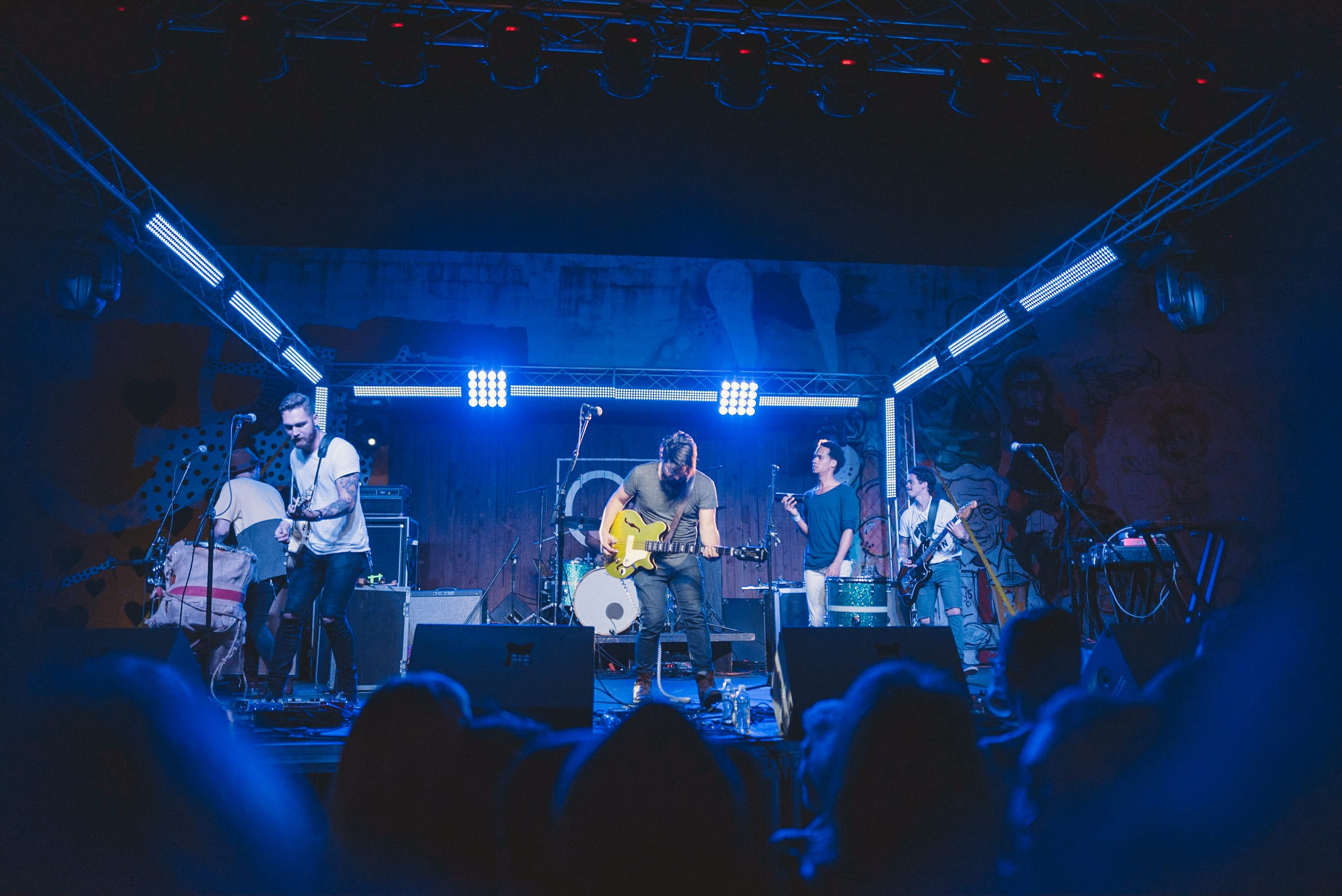 Male guitarist with black hair and beard jamming with yellow guitar with fellow male musicians and guitarists around him.