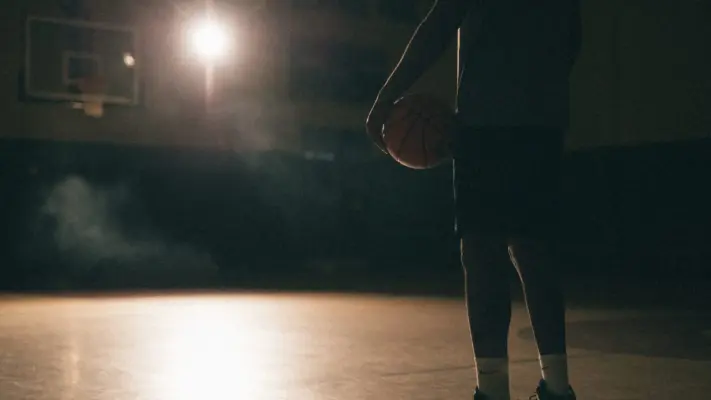 Corporate and Commercial Video Production 1 Side profile of the lower half of an African American man poised holding a basketball on a basketball court with a light shining on him in semi darkness Nike Sneakers