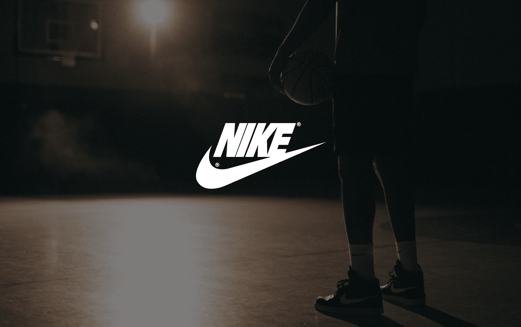 IU C&I Studios Page Nike Sneakers Conceptual Advertisement White Nike logo with a background showing the lower half of an African American man poised holding a basketball on a basketball court with a light shining on him in semi darkness