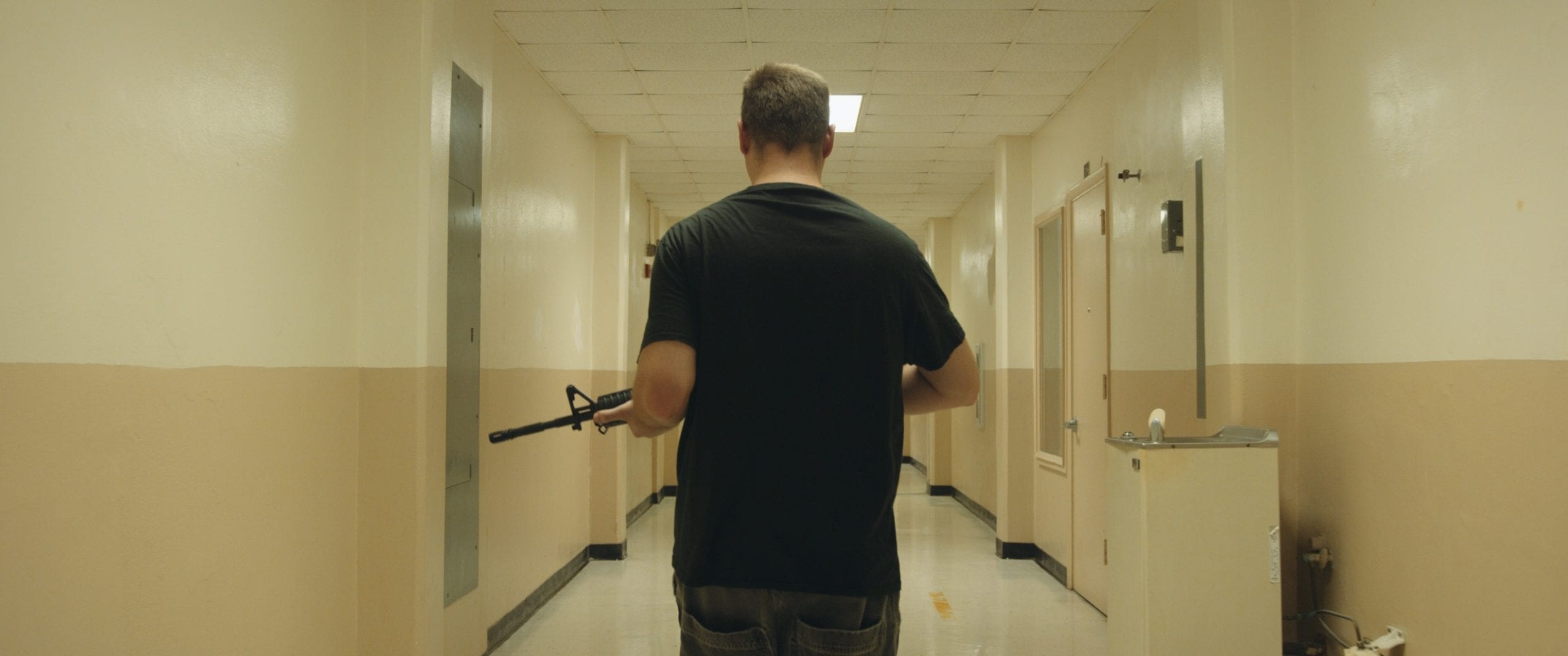 Black Violin One Step Music Video View from behind of a man wearing a black t shirt walking down a business corridor holding an assault rifle