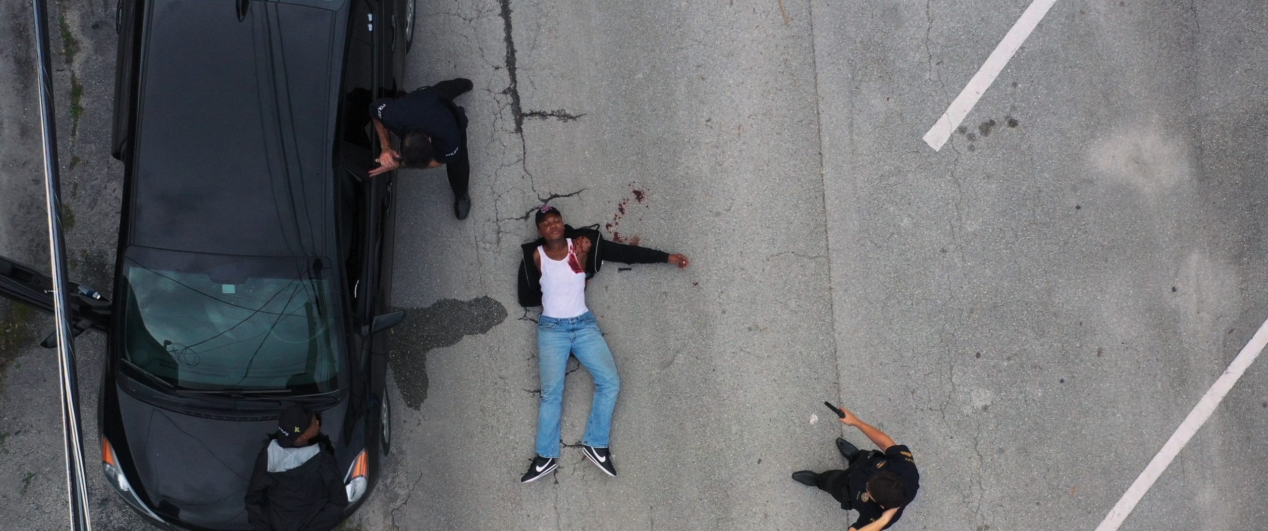 Black Violin One Step Music Video Overhead view of two police officers by a black car and a man wearing white tank top and jeans laying shot on the street