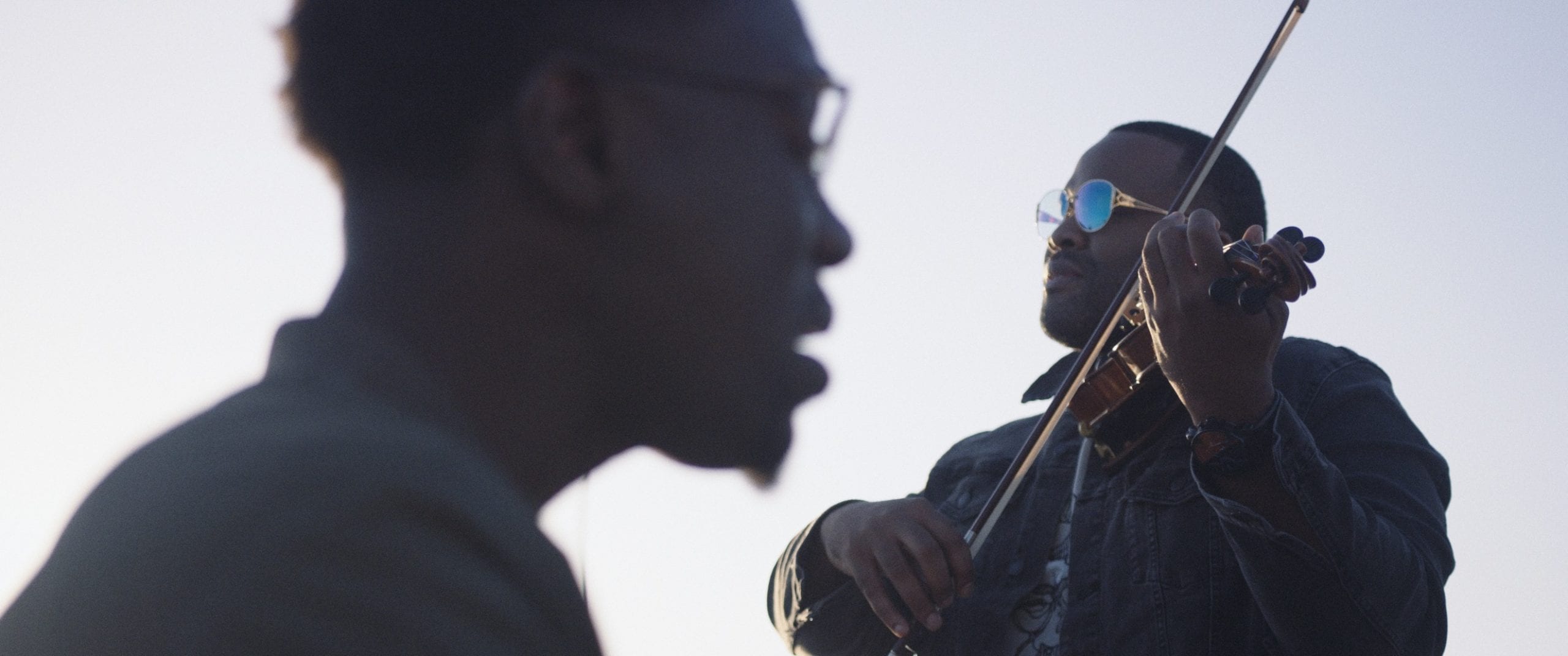 Black Violin One Step Music Video Side profile headshot of a man wearing glasses and another man wearing glasses playing a violin both posing for the camera