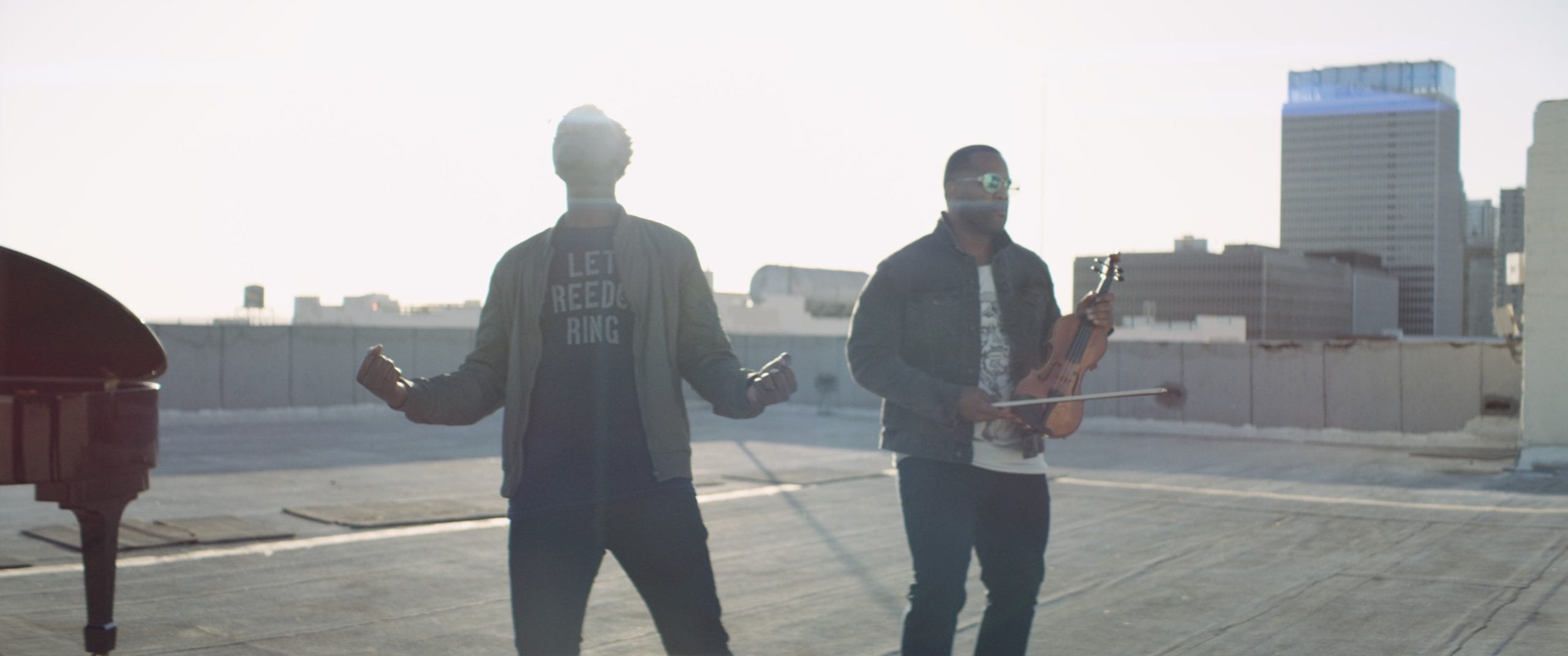 Black Violin One Step Music Video Two men by a piano posing for the camera with one looking at the sky and the other holding a violin on a concrete rooftop