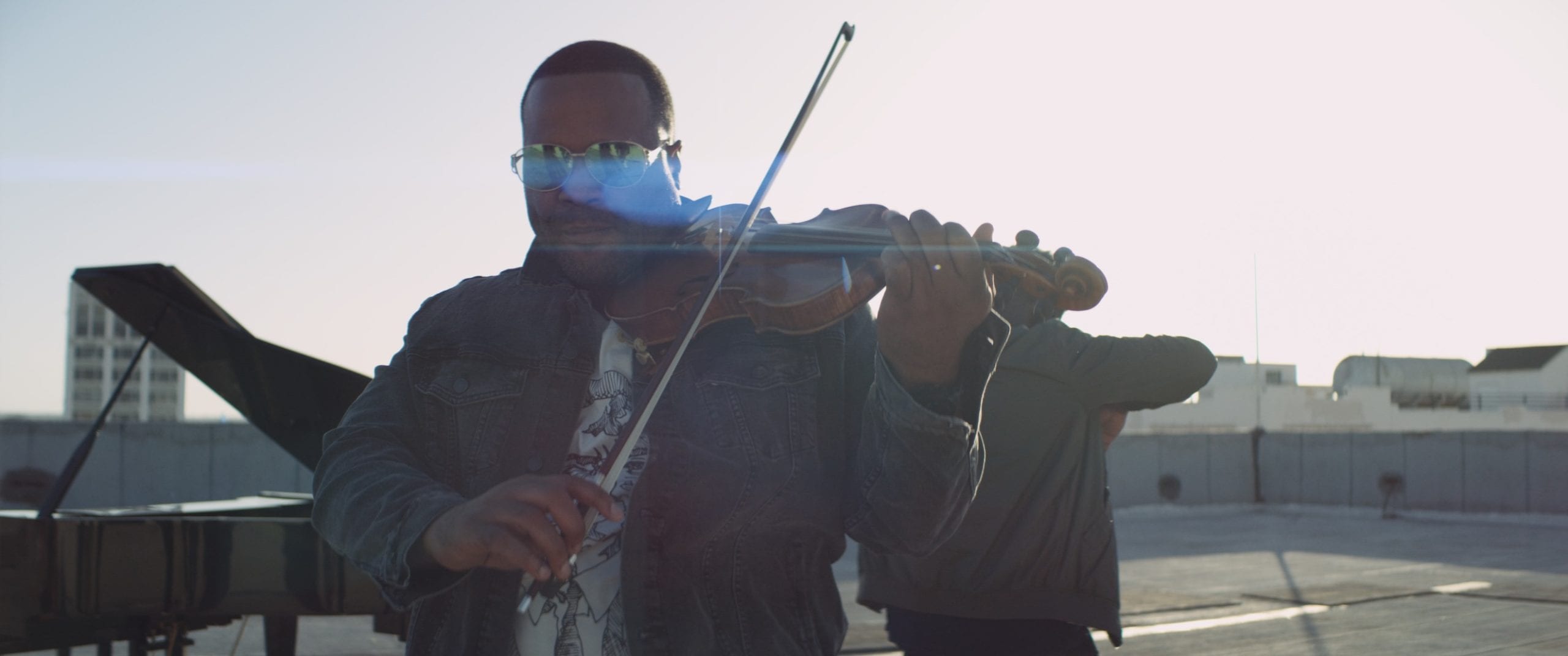 Black Violin One Step Music Video Closeup of man wearing shades playing a violin on a concrete rooftop with a piano nearby
