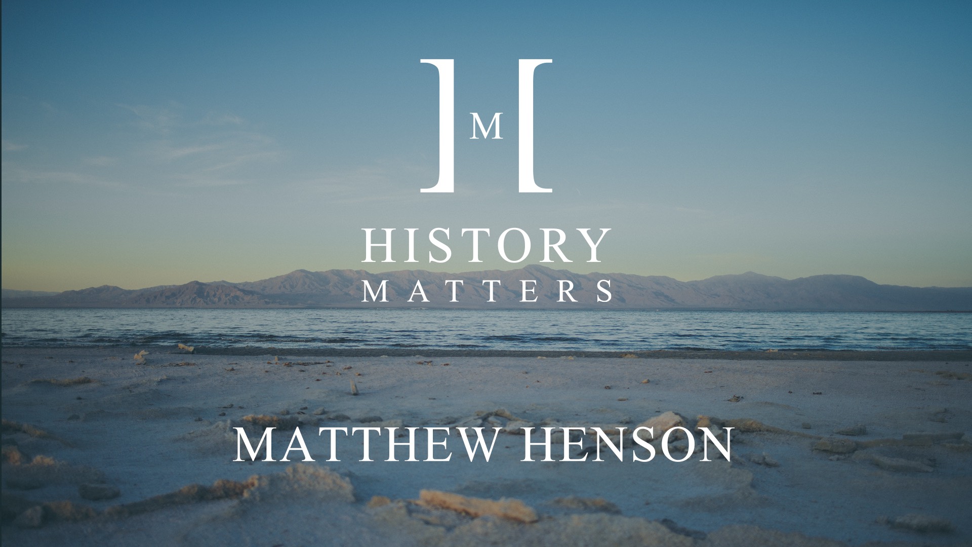 White HM Matthew Henson logo with background view of sand and beach with mountains in the background