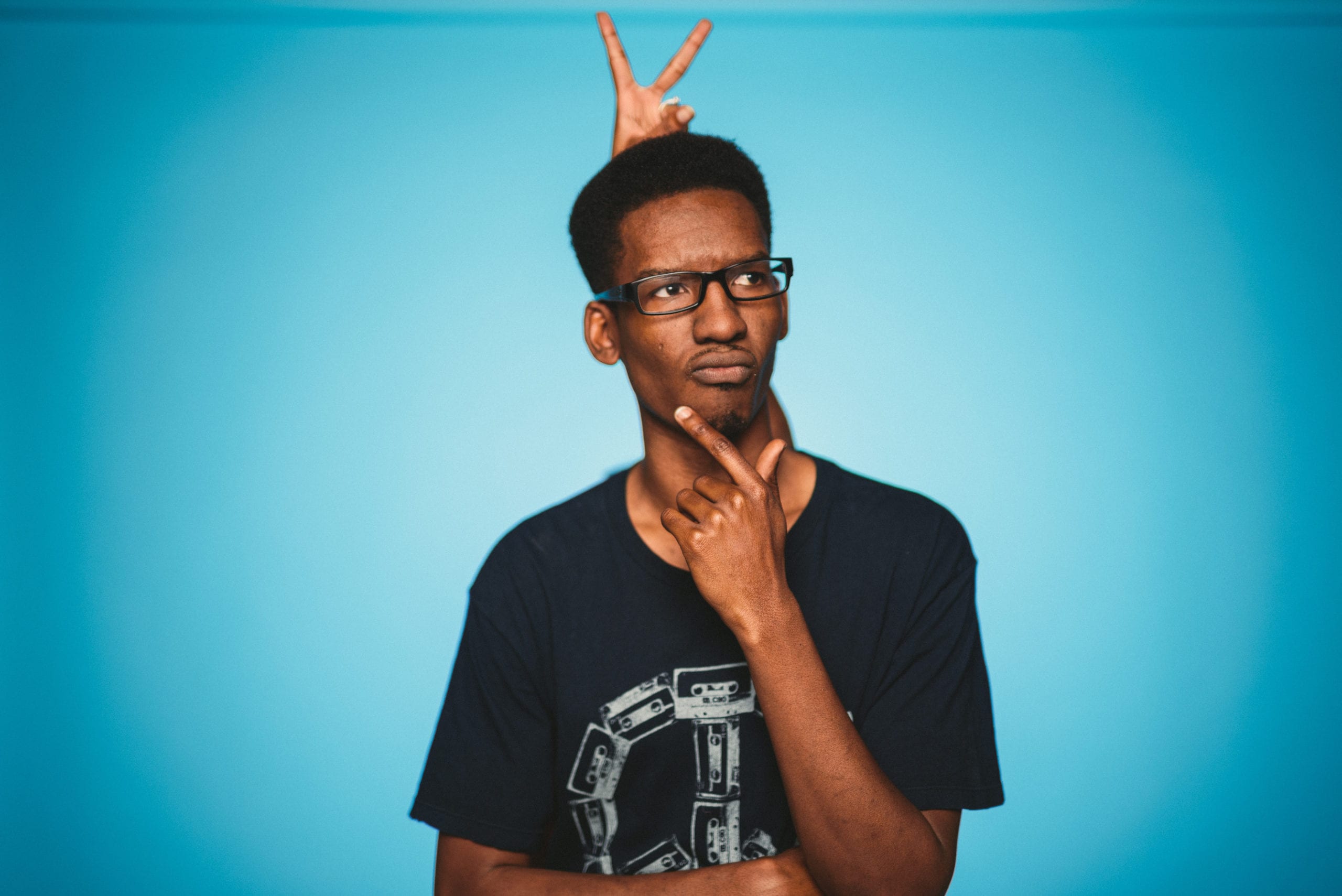 IU C&I Studios Portfolio Heart Piece Media Day African American man doing a thinking pose for the camera in a light blue light in a black t shirt with someone doing rabbit ears behind him