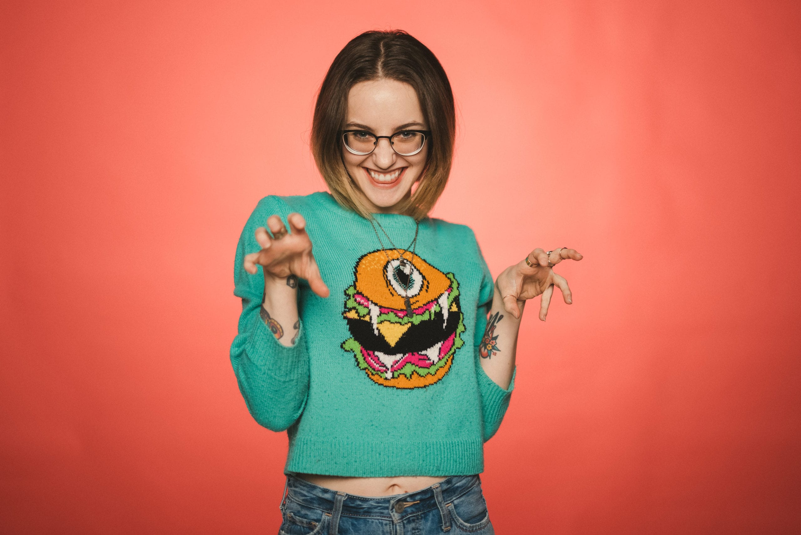 Heart Piece Media Day Woman with short brown hair wearing glasses posing with clawed hands wearing a turquoise sweater with a graphic of a hamburger with an eye on it