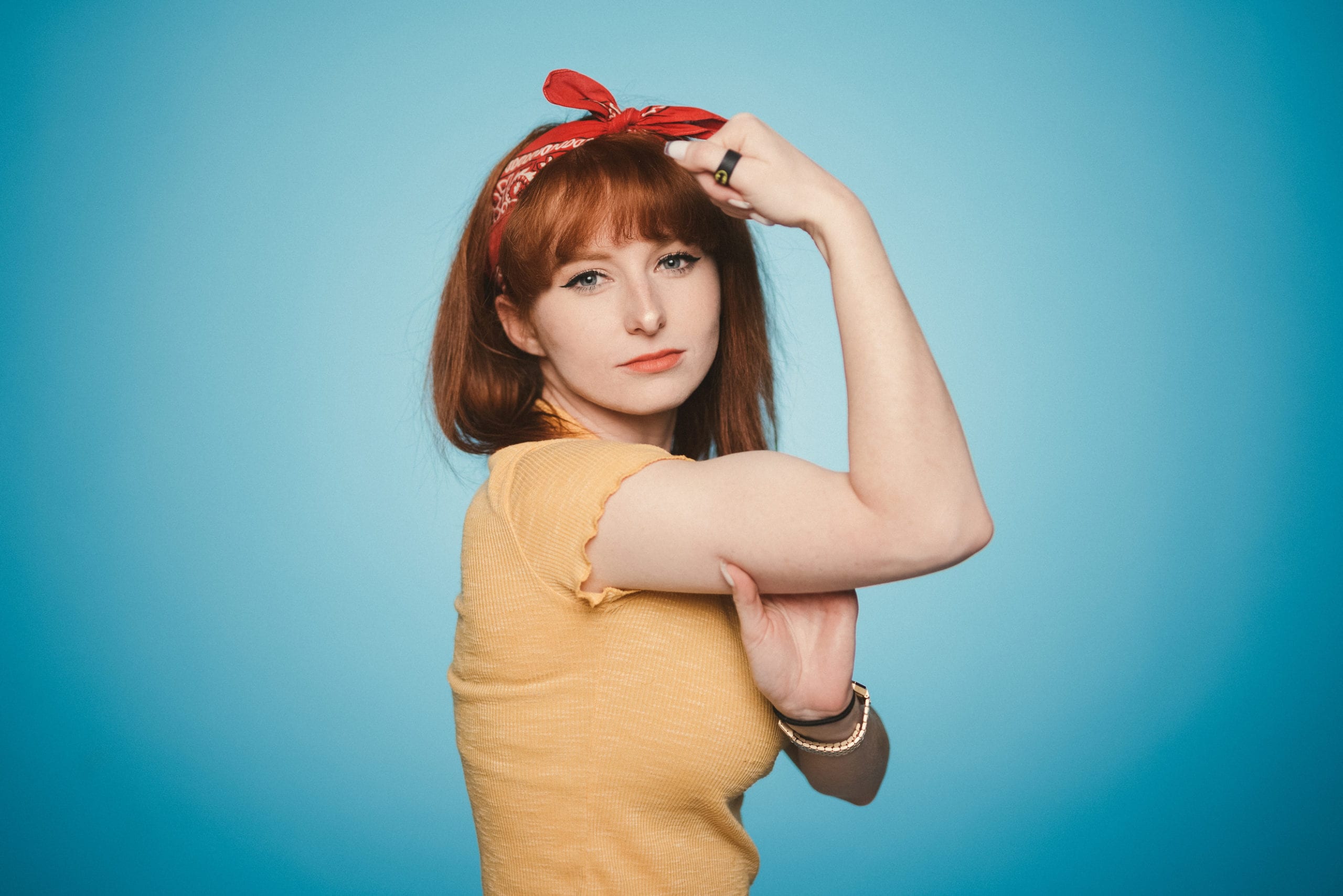 IU C&I Studios Portfolio Heart Piece Media Day Woman with short hazel hair with a red ribbon in it doing a Rosie the Riveter pose for the camera with a light blue background