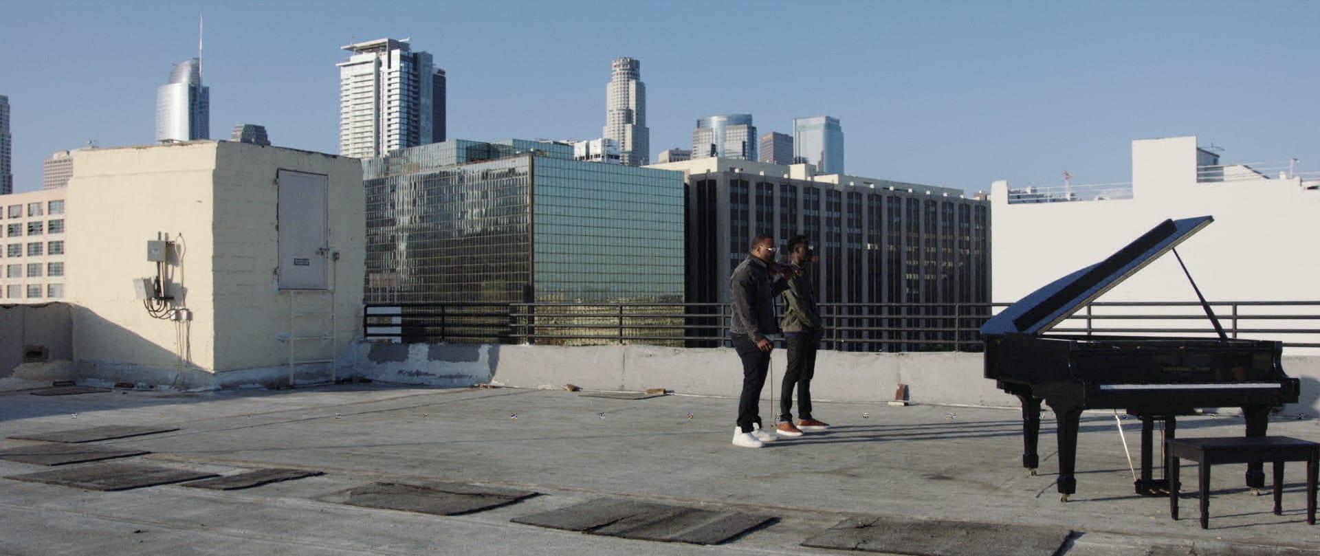 One Step Music Video Black Violin Side view of two men wearing glasses standing by a piano on a concrete rooftop