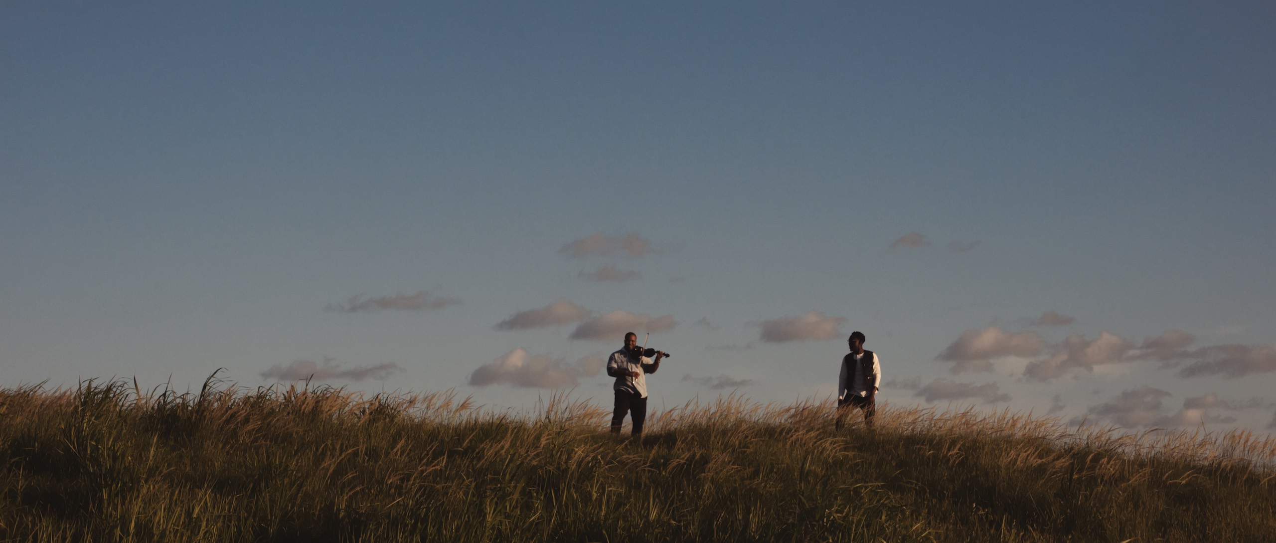 Black Violin Impossible is Possible Music Video with man playing violin in a field under cloudy blue skies as another man looks on