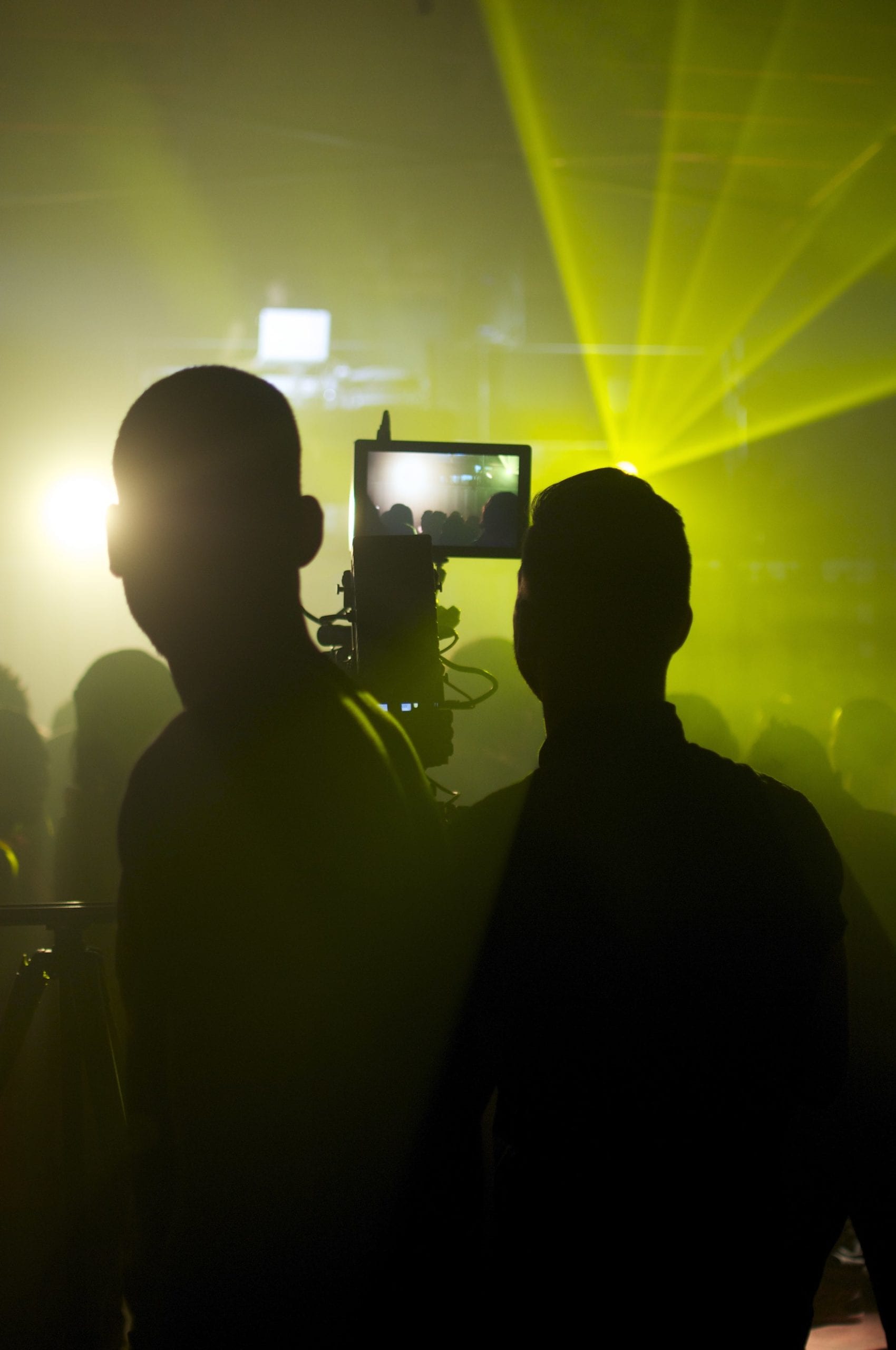 View from behind silhouette of two video camera operators filming a concert basked in yellow light