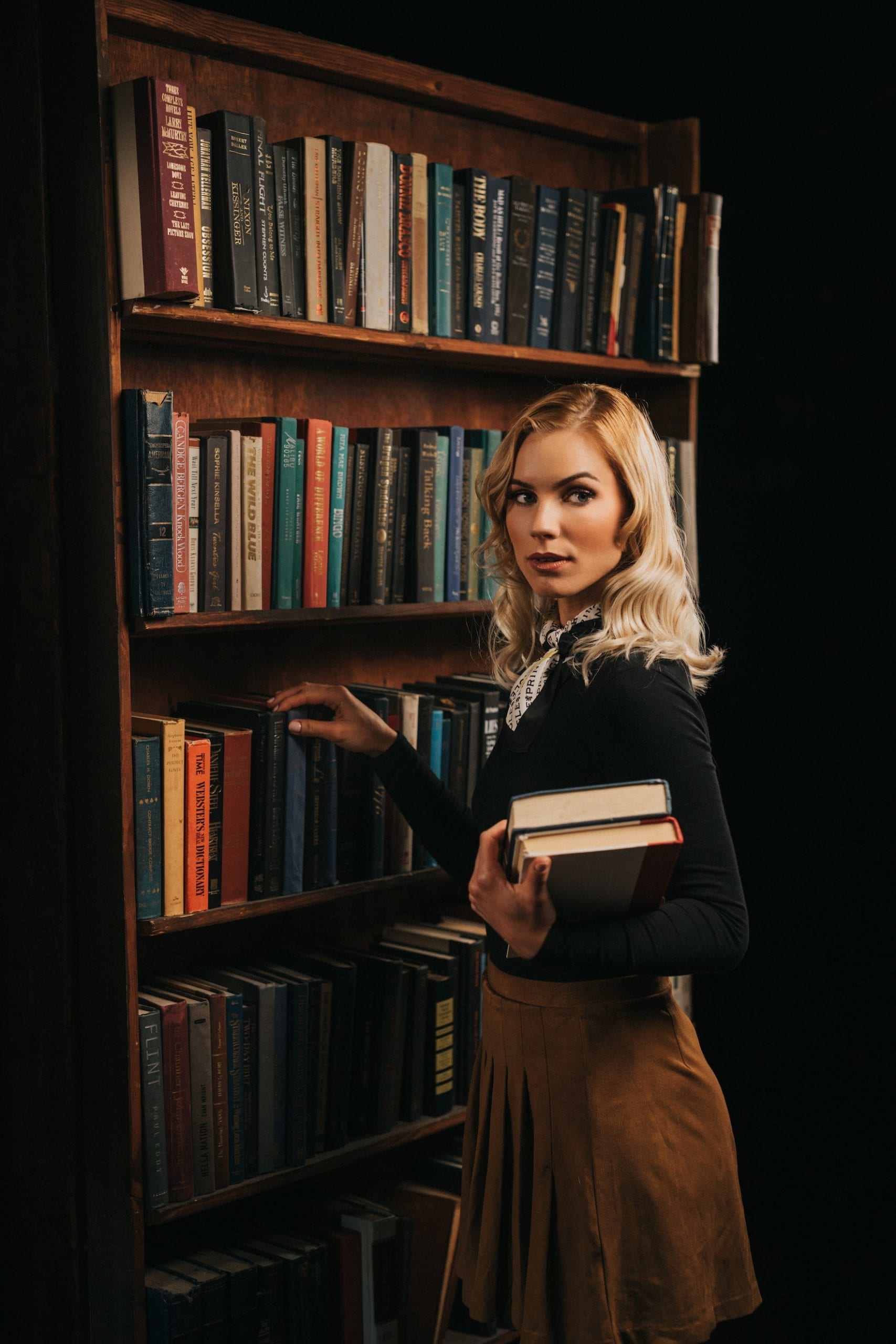 Professional Photography Services by C&I Studios Woman with long blond hair posing by a bookcase with books and holding two books in her arm