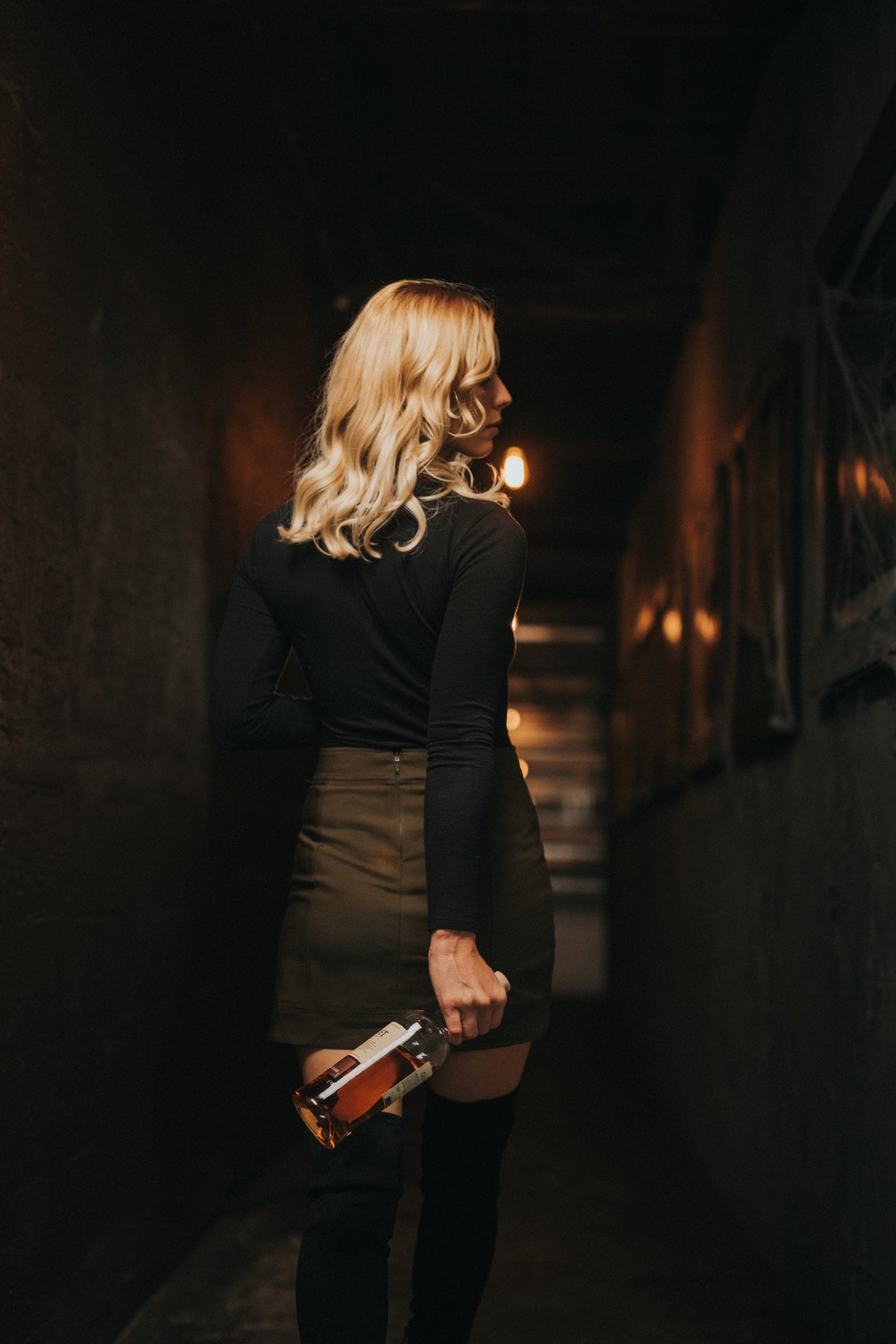 View from behind of woman with long blond hair walking carrying a liquor bottle looking off to the side