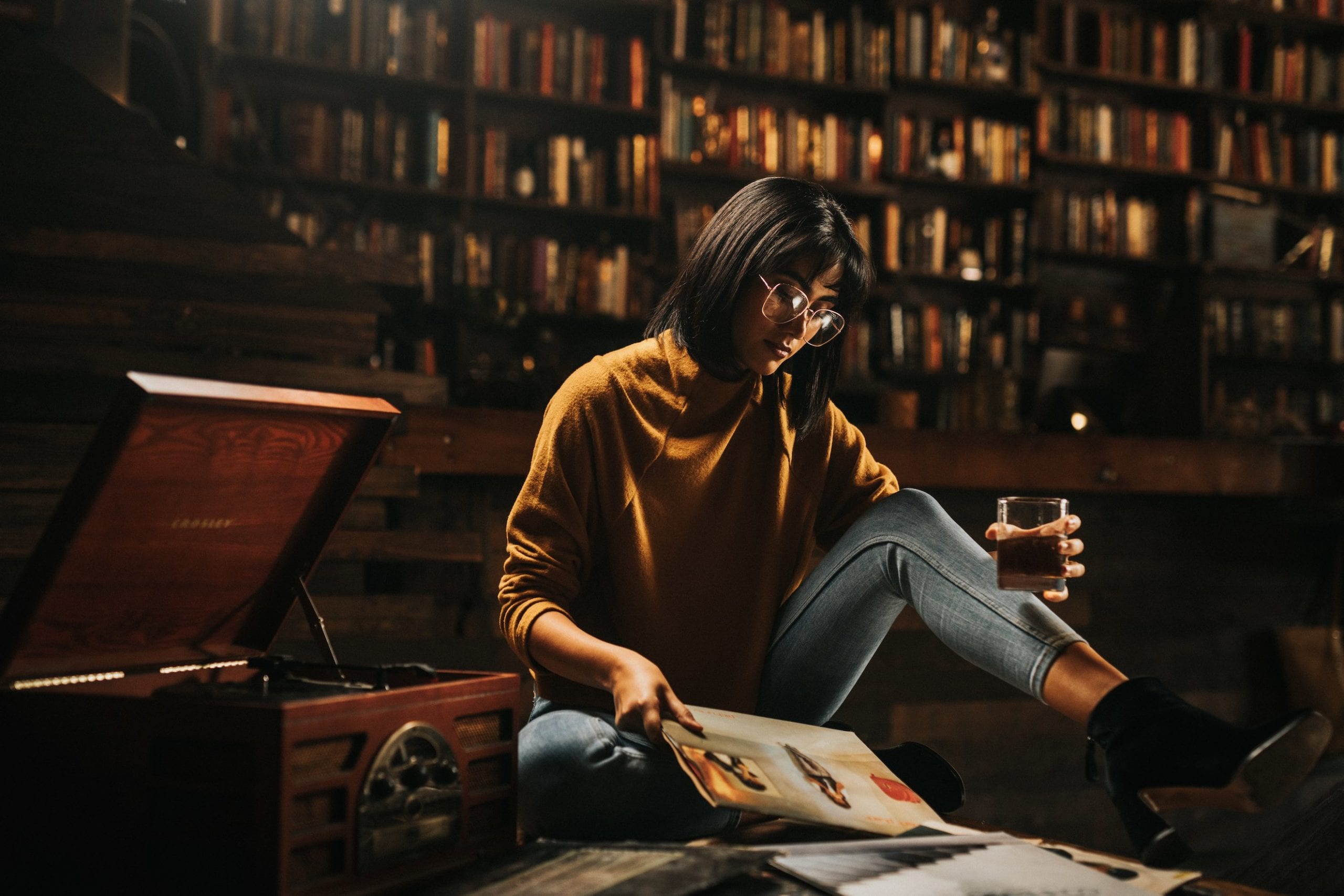 Woman posing for camera wearing glasses, jeans and burnt orange top holding a glass drink looking down at ads next to an old vinyl record player