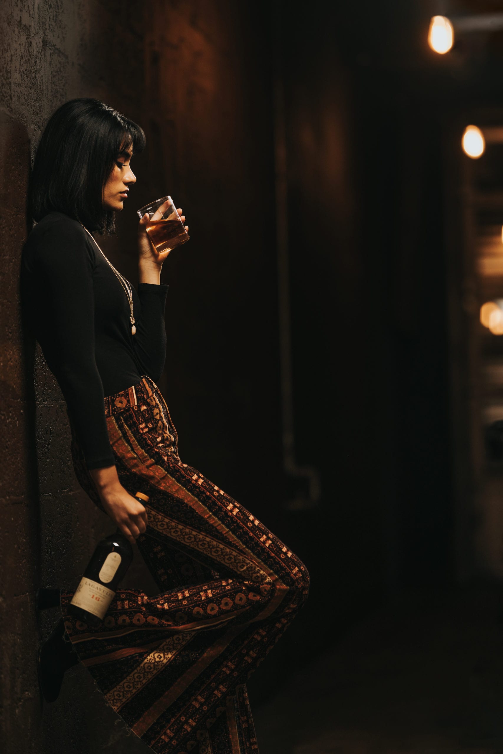IU C&I Studios Portfolio Next Door Rebranding and hospitality photoshoot Side profile of woman posing for camera with short black hair wearing black turtleneck and patterned pants leaning against the wall holding a glass drink in one hand and an bottle of alcohol in the other