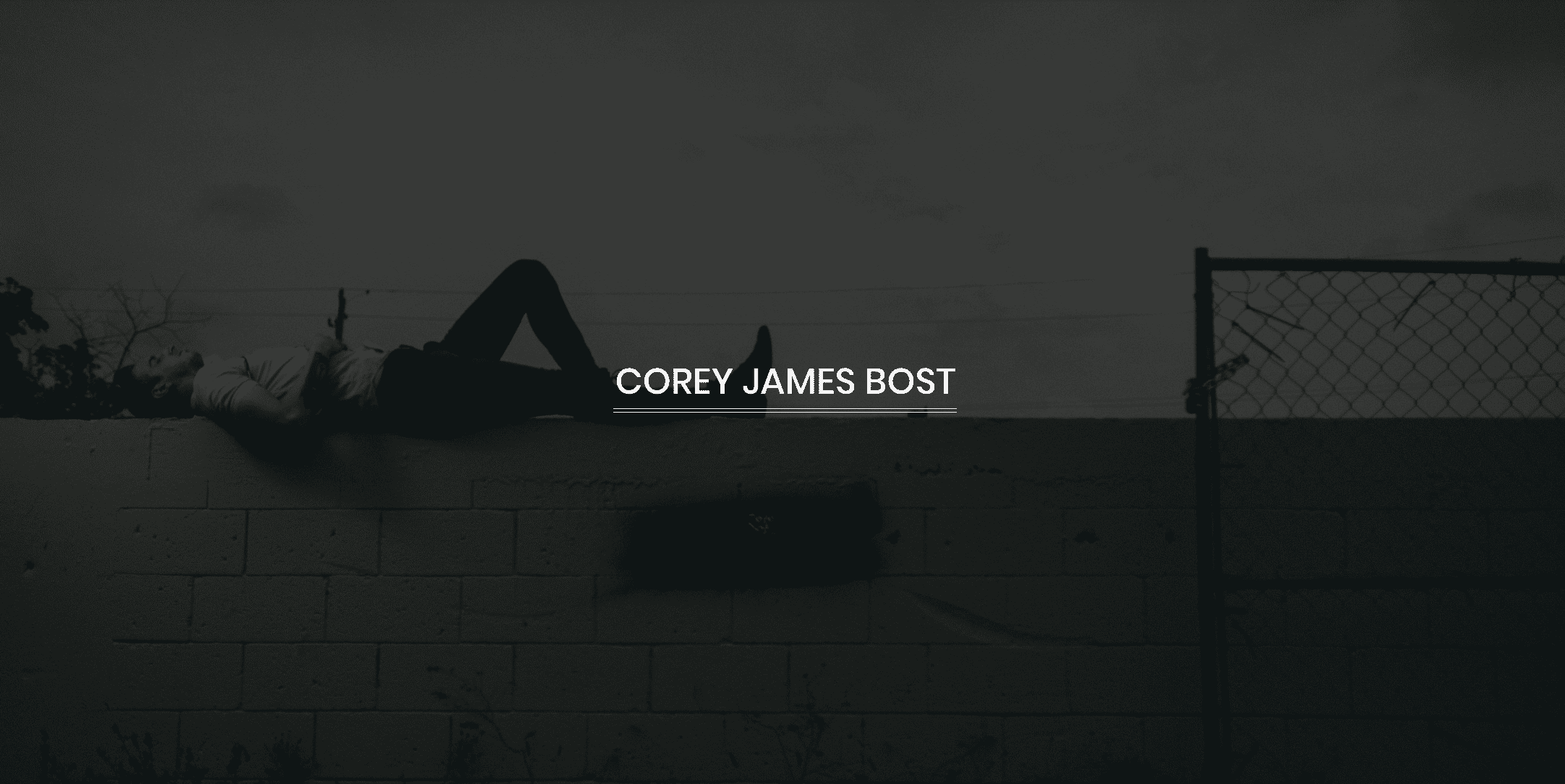 White Corey James Bost logo with black and white background of tattooed man posing for the camera laying on a wall
