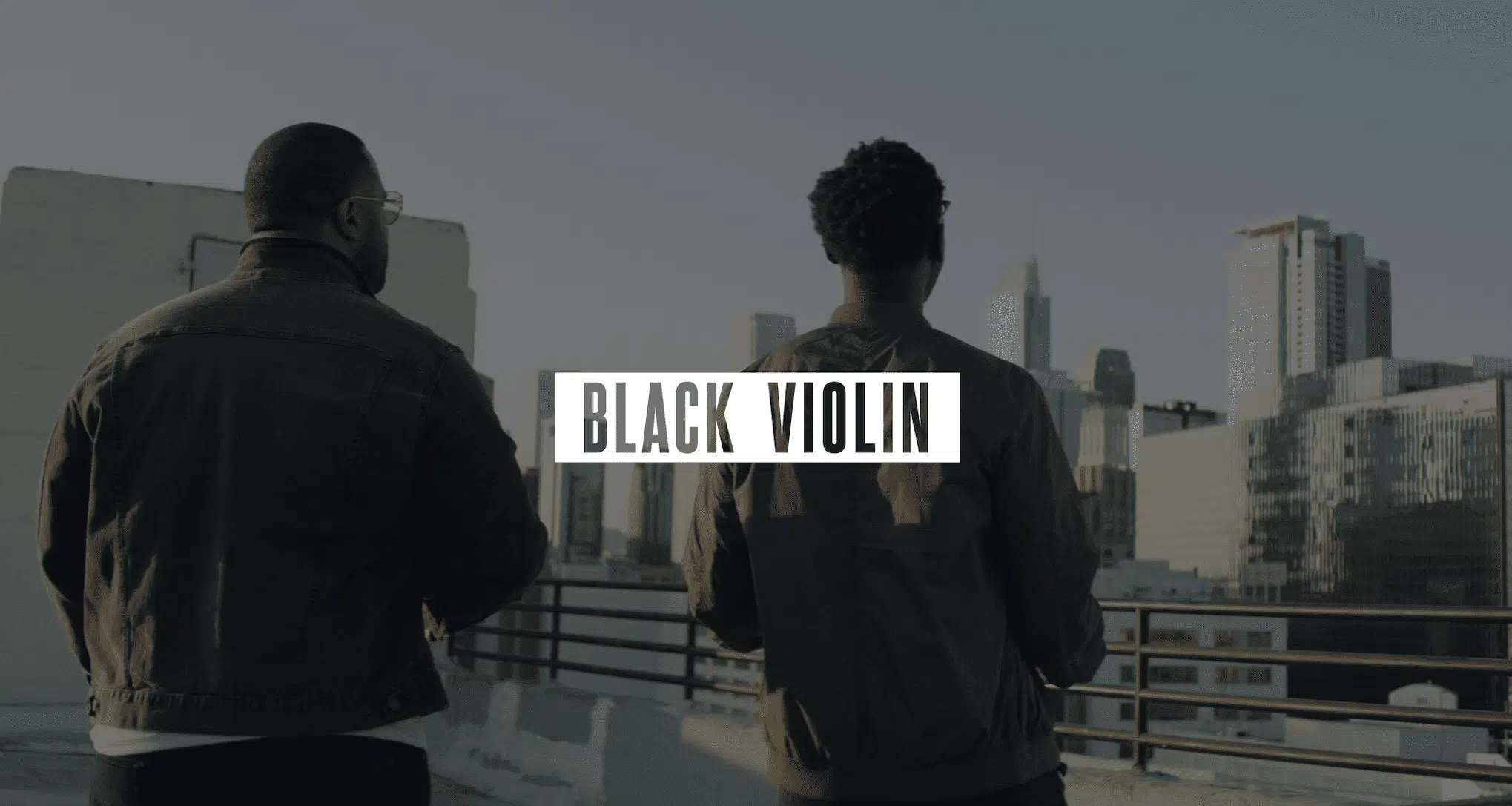 Keyword: Artist Management Services .Video Production Services to Podcast Recording Studio Gray Black Violin logo on white against a dimmed background of two men standing on a concrete rooftop looking out over the city