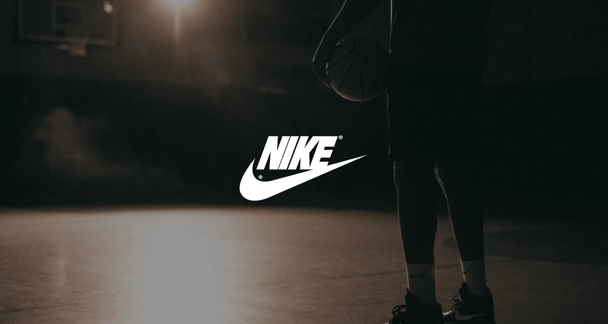 White Nike logo against a dim view from behind background of a man holding a basketball on a basketball court. Video Production Services