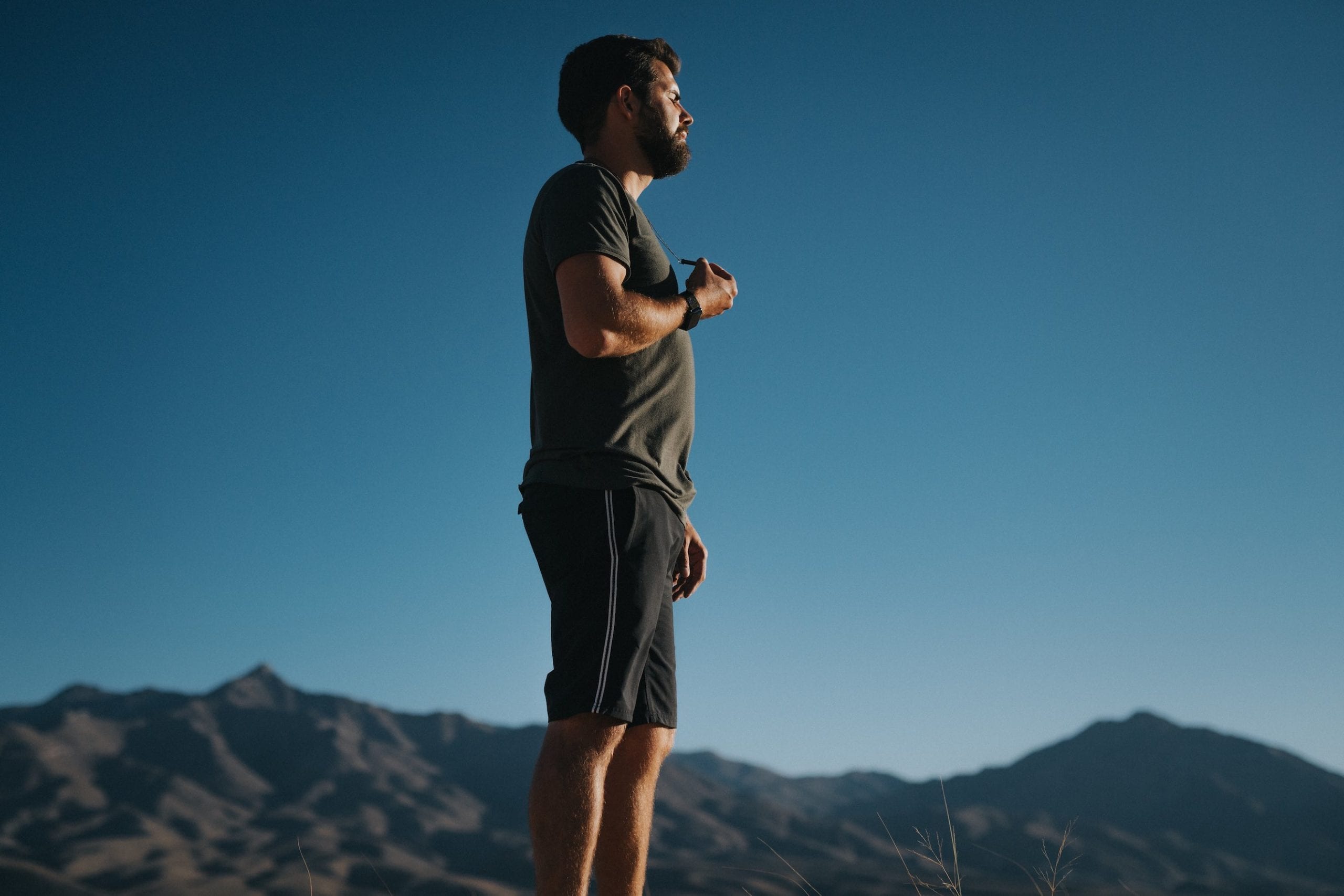 Side profile of a man with short hair and beard wearing a gray t shirt and black shorts posing for the camera with eyes closed with hills in the background