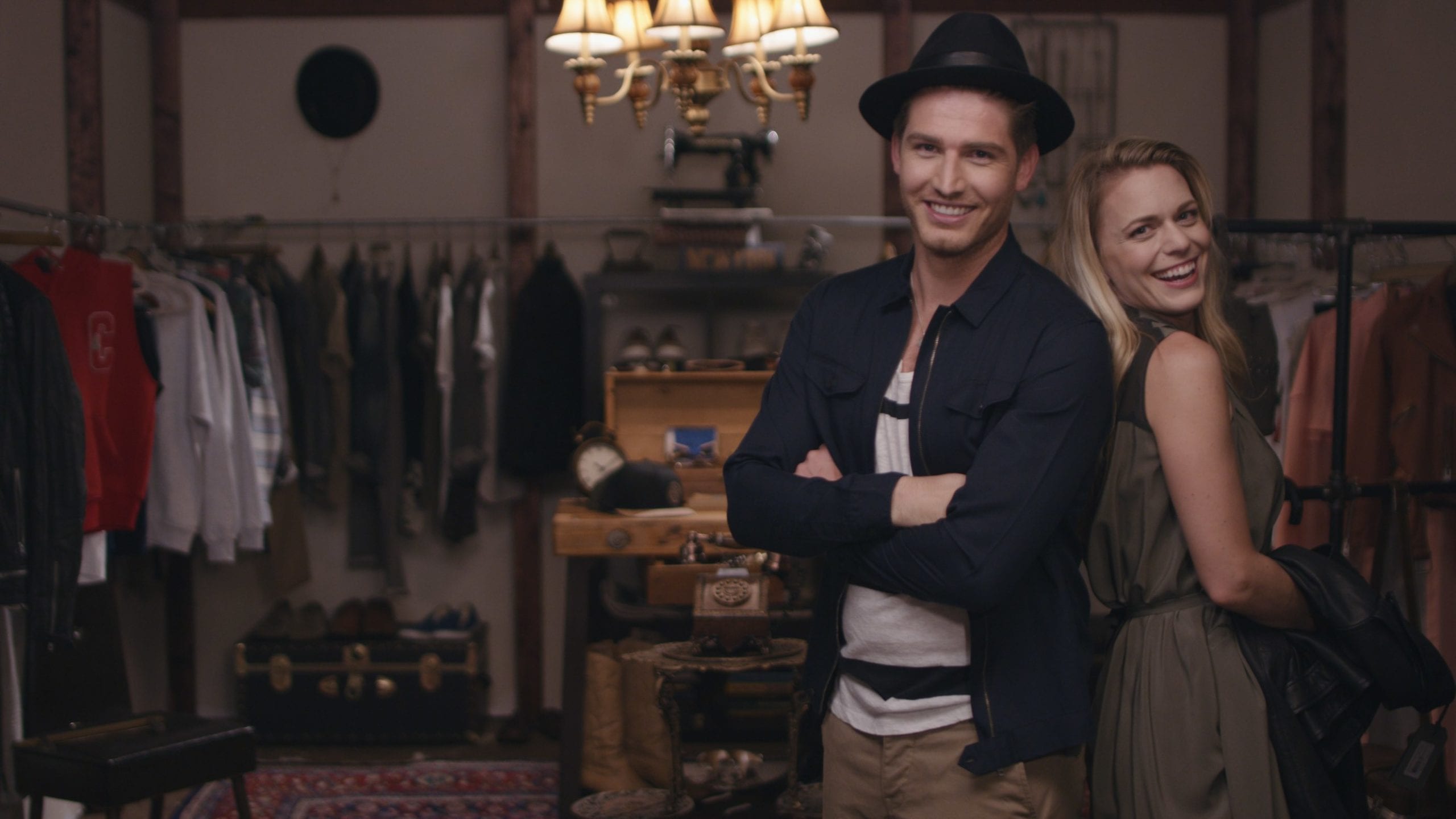 AllSaints Clothing Brand Man wearing a black hat and dark blue shirt standing with his back against a woman wearing a dress and long blond hair in a wardrobe room smiling and posing for the camera