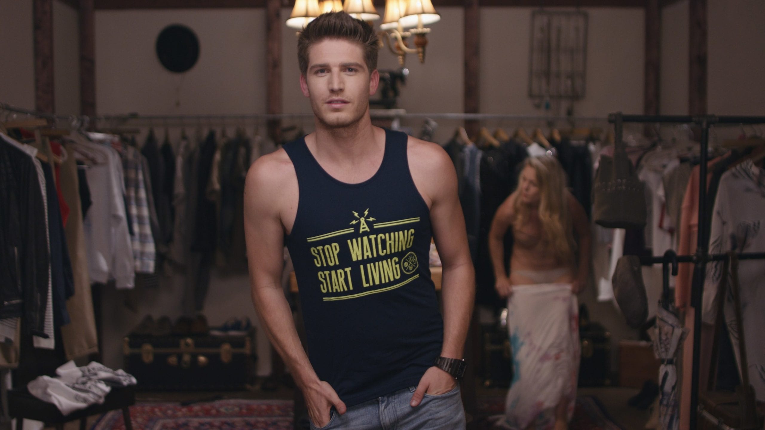 AllSaints Clothing Brand Young man with short hair wearing a black tank top that says Stop Watching Start Living in a wardrobe room with a woman putting on a dress in the background